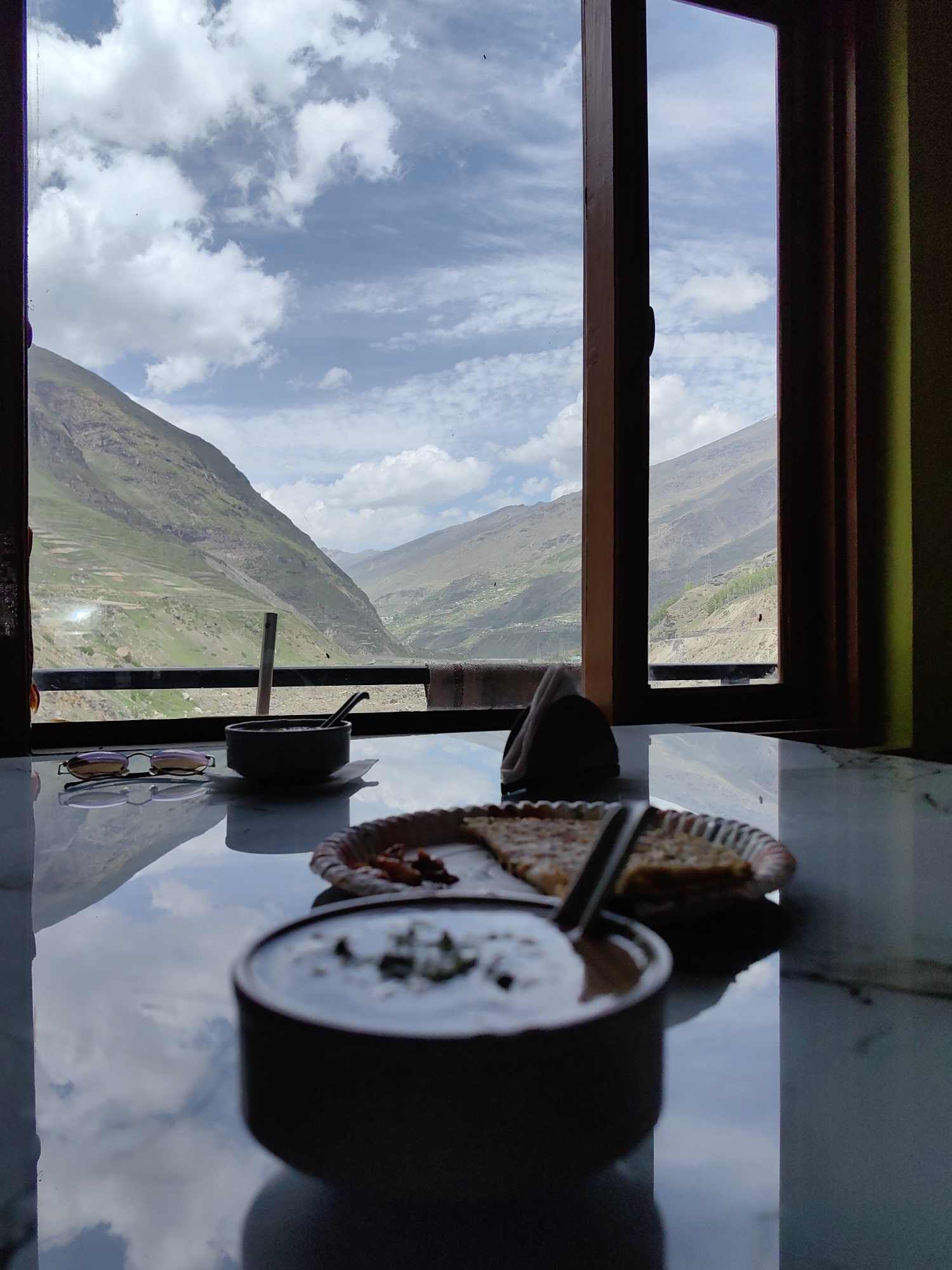 The image depicts a scenic view from a window overlooking the mountains and valleys surrounding Sissu, India. In the foreground, on a reflective marble-like table, is a meal consisting of a bowl of yogurt or raita with herbs, and a plate of what appears to be flatbread or paratha with a side of red sauce. To the left of these food items, a pair of sunglasses rests on the table. The window frame, made of dark wood, divides the view of the landscape. The exterior view shows lush green hillsides, dotted with some structures and trees, under a bright sky filled with white, puffy clouds. The reflection on the table captures the sky and the surrounding environment, enhancing the sense of depth and immersion. The overall scene suggests a tranquil dining experience with a breathtaking natural backdrop.