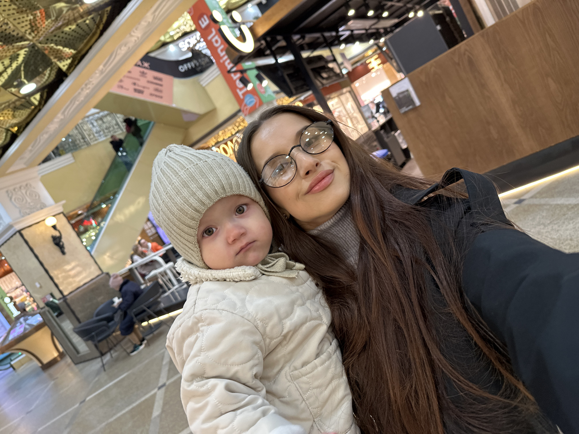 A woman with long brown hair and glasses is holding a toddler in a beige hat and jacket. They are taking a selfie in a shopping mall in Yekaterinburg, Russia. The mall has multiple levels with shops and seating areas. The ceiling has a mirrored design.