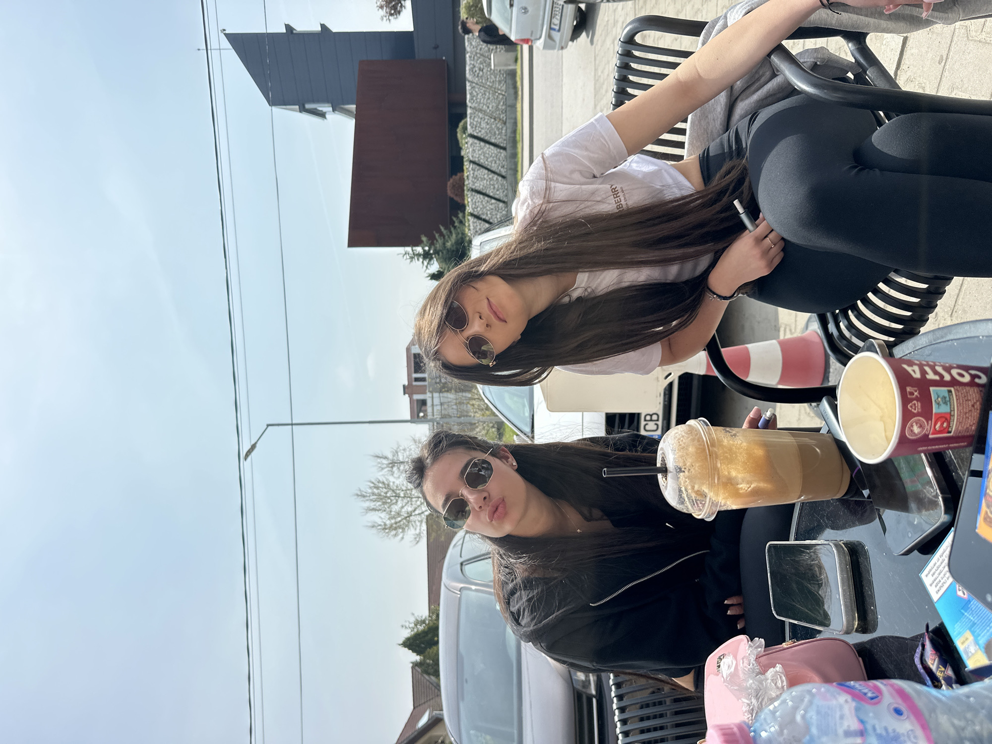 Two young women are posing outdoors at a cafe in Plovdiv, Bulgaria. One is wearing a white t-shirt and black leggings, sitting on a metal chair. The second is wearing a black jacket and sunglasses, seated at a black table with a plastic cup of iced coffee, a Costa coffee cup, mobile phones, a pink handbag, and a water bottle. Cars and buildings are visible in the background. The weather appears sunny.