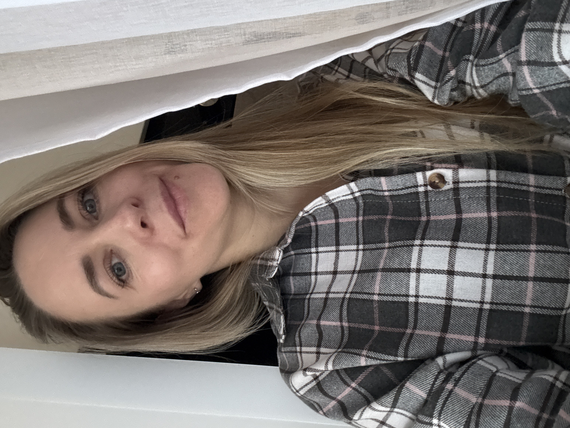 A blonde woman is shown in a close-up, slightly rotated portrait. She has light skin, blue eyes, and is wearing a gray, white, and pink plaid flannel shirt. She is positioned close to a white curtain or fabric. The background and specific location in Chaikovsky, Russia, are not discernible.