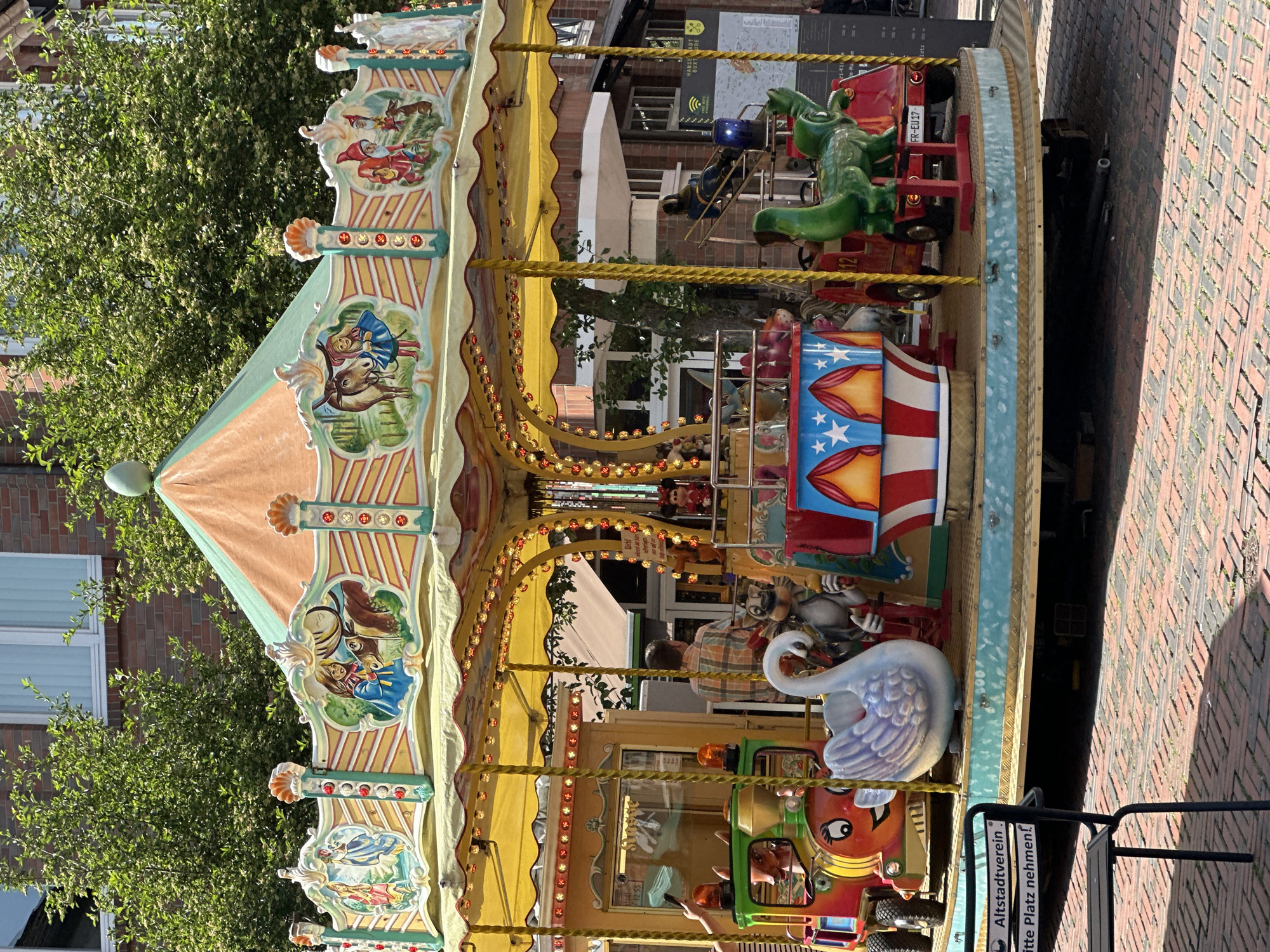 The image shows a vintage carousel in Buxtehude, Germany. The carousel has a tent-like top with illustrations of fairy tale characters and landscapes. The base is decorated with lights and various ride vehicles, including a swan, a train, and a green dinosaur. The surrounding environment includes a brick street, buildings, trees, and a sign that says "Alstadtverein bitte Platz nehmen!"