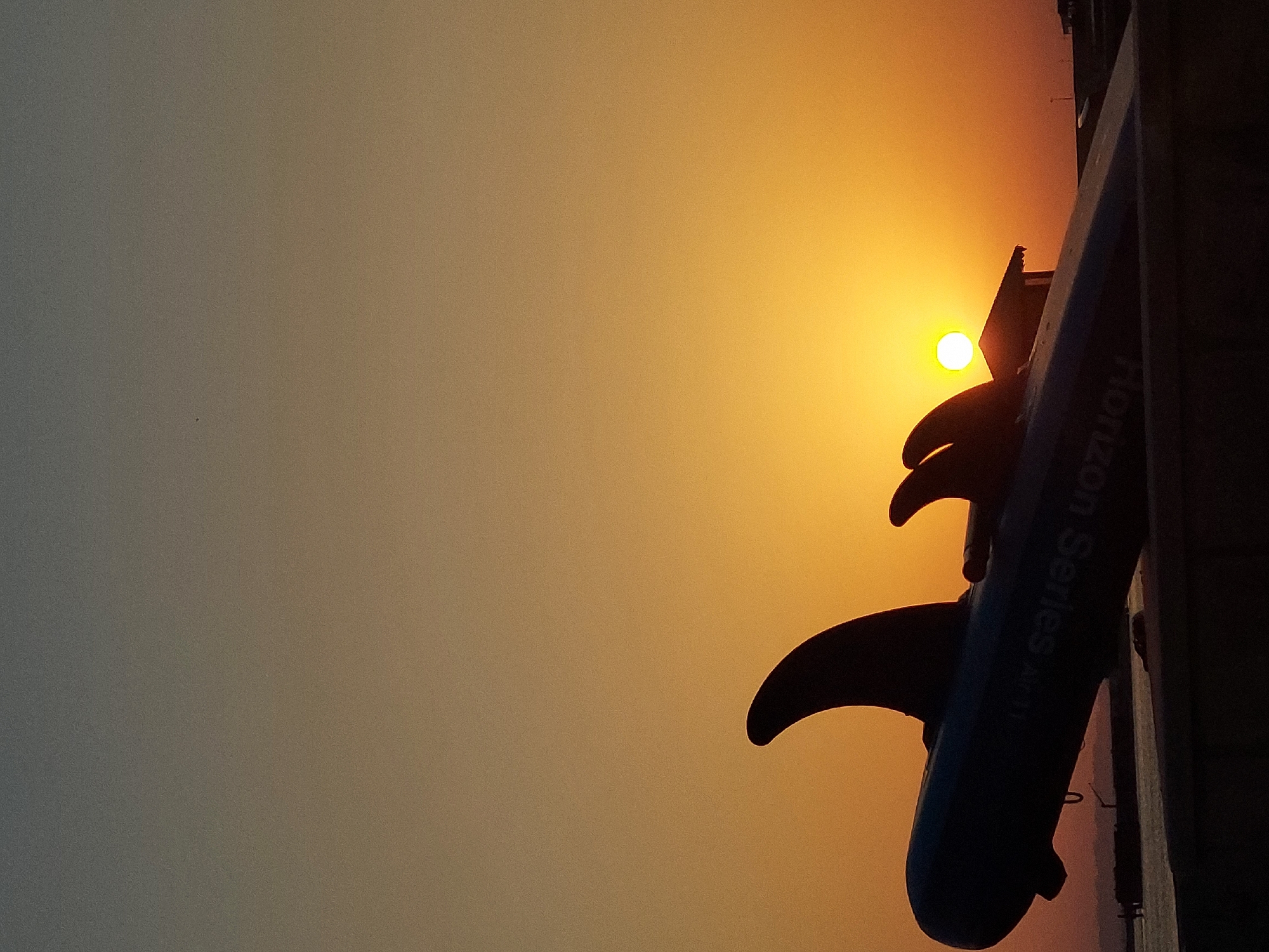 An inflatable dolphin-shaped toy is positioned on a concrete barrier at sunset in the city of Pirallahi Raion, Azerbaijan. The toy is predominantly blue and features two fins, one of which is prominently displayed in the foreground, silhouetted against the warm hues of the setting sun. The sun itself appears as a bright, yellow orb, slightly above the horizon, casting a golden glow across the sky. In the background, a small, dark structure with a triangular roof is visible, partially obscured by the toy. Further in the distance, the silhouette of a large cargo ship and industrial cranes can be seen against the fading light. The sky transitions from a deep grey at the top to a vibrant orange and yellow near the horizon. The overall scene is one of a serene evening by the water.