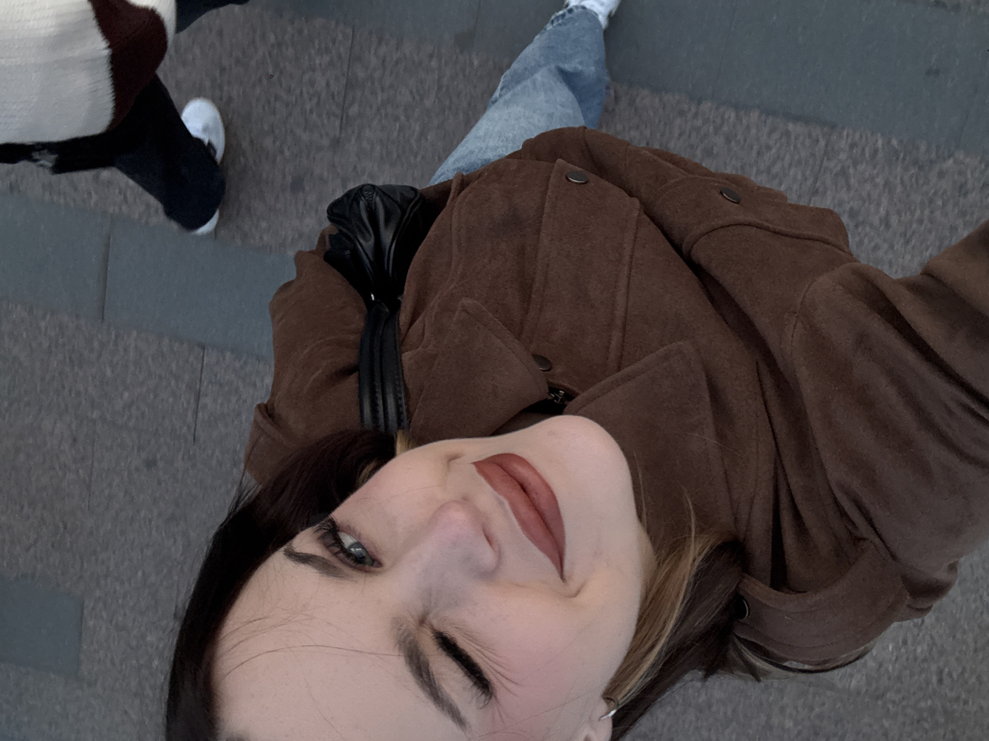 A high-angle, medium shot captures a young woman taking a selfie outdoors on a grey, tiled surface. She is wearing a brown jacket and blue jeans. Her right eye is closed in a wink, and her left eye is open and looking at the camera. Her lips are painted a muted red. A black purse is slung over her shoulder. In the background, another person's legs and feet, clad in white sneakers, are visible. The lighting suggests it is daytime. There is no discernible landmark that would indicate Chelyabinsk, Russia.
