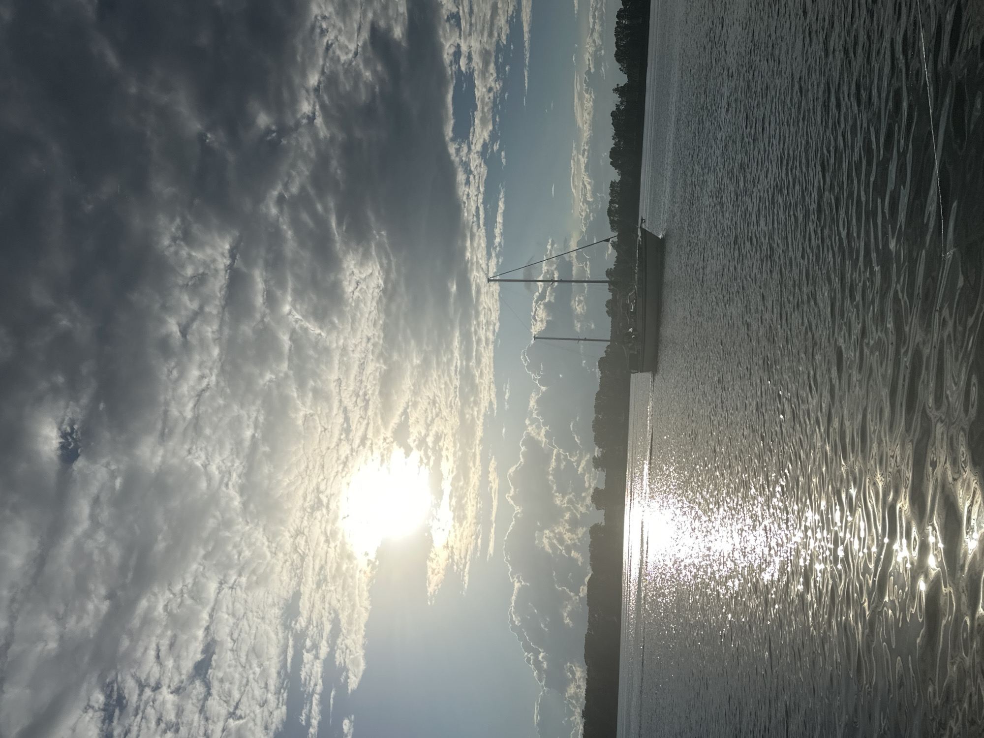 A sailboat is docked on the water near a wooded shoreline. The sun is low in the sky, casting a golden glow on the water and clouds. The clouds are dramatic and textured, with some appearing dark and heavy, while others are wispy and illuminated by the sun. The water is choppy, with sunlight reflecting off the waves. The scene suggests a calm and serene moment, possibly at sunrise or sunset. The orientation of the image is vertical.
