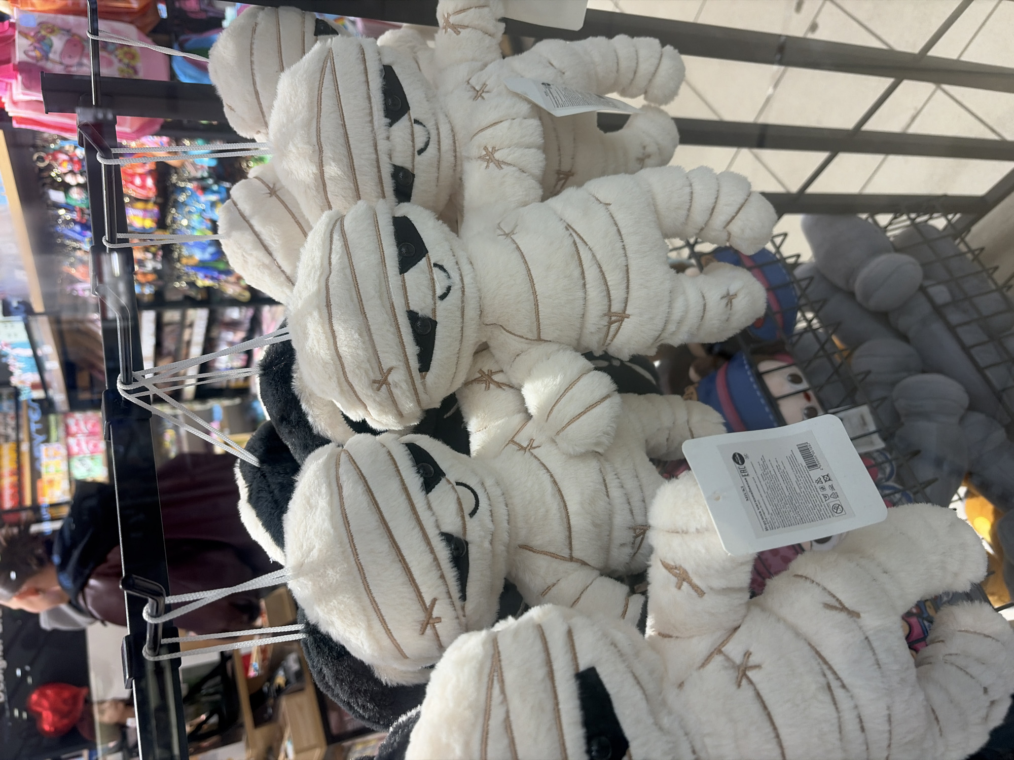 This photograph shows a display of white, plush toy mummies in a store. The toys are hanging from a rack and are arranged in a pile. The location is Saint Petersburg, Russia, as indicated by the product tags visible in the photo. A person is visible in the reflection of the store's display window in the background. Other merchandise is visible on the shelves and racks behind the plush toys. The environment appears to be a retail setting, likely a shop selling souvenirs or gifts.