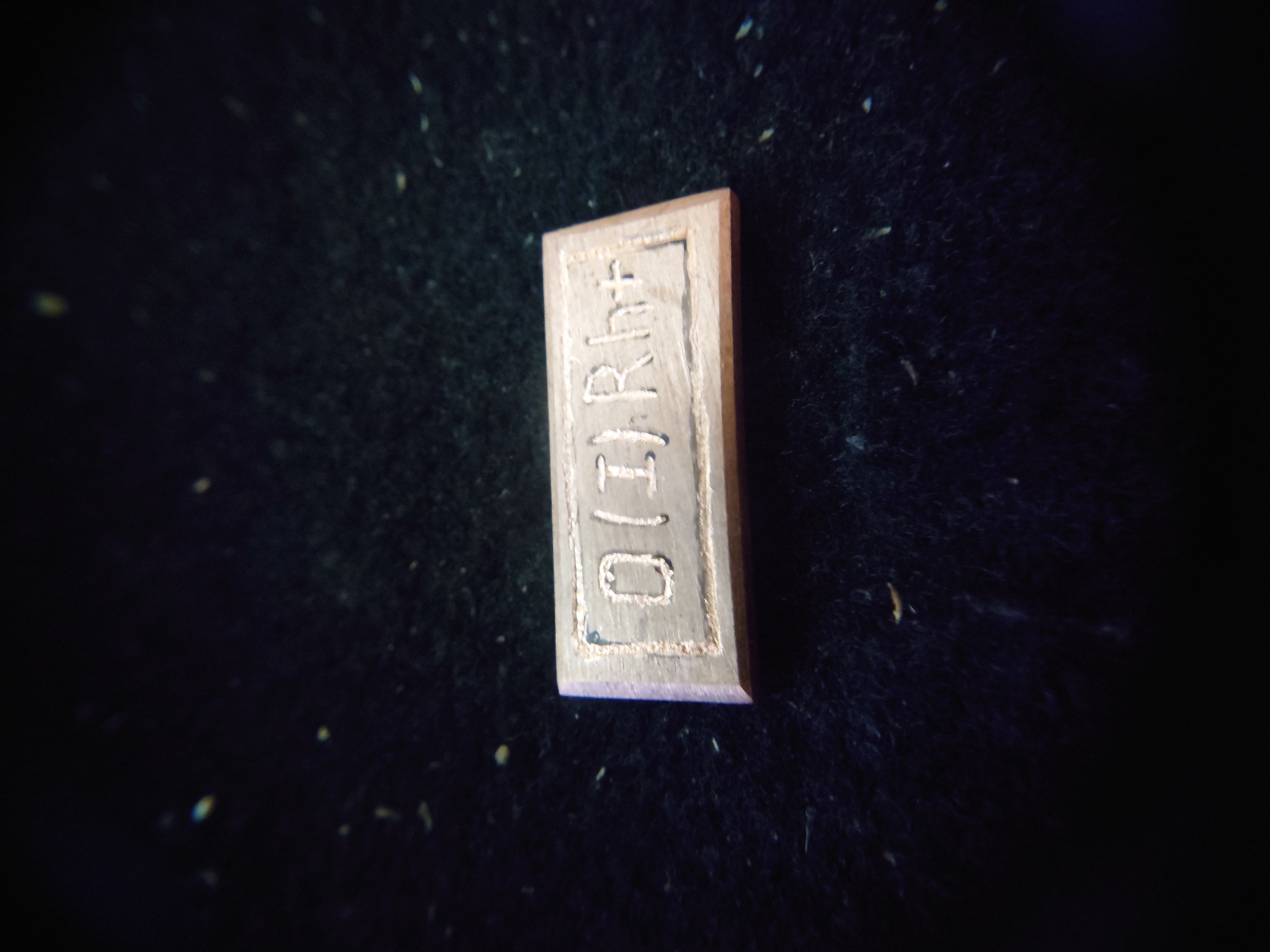 A rectangular metal tag with the inscription "0(I)Rht" is placed on a dark, textured surface. The tag is made of a light-colored metal, possibly copper or brass, and is slightly beveled on its edges. The inscription appears to be etched or stamped into the surface of the tag. The dark background is out of focus and appears to be fabric or a similar material, with small specks scattered across it. The lighting is dim, casting a slight shadow on the surface beneath the tag. The object is centrally located in the frame. There are no people or other objects visible in the image. There is no information that can place this object in Sokolohirsk, Ukraine.