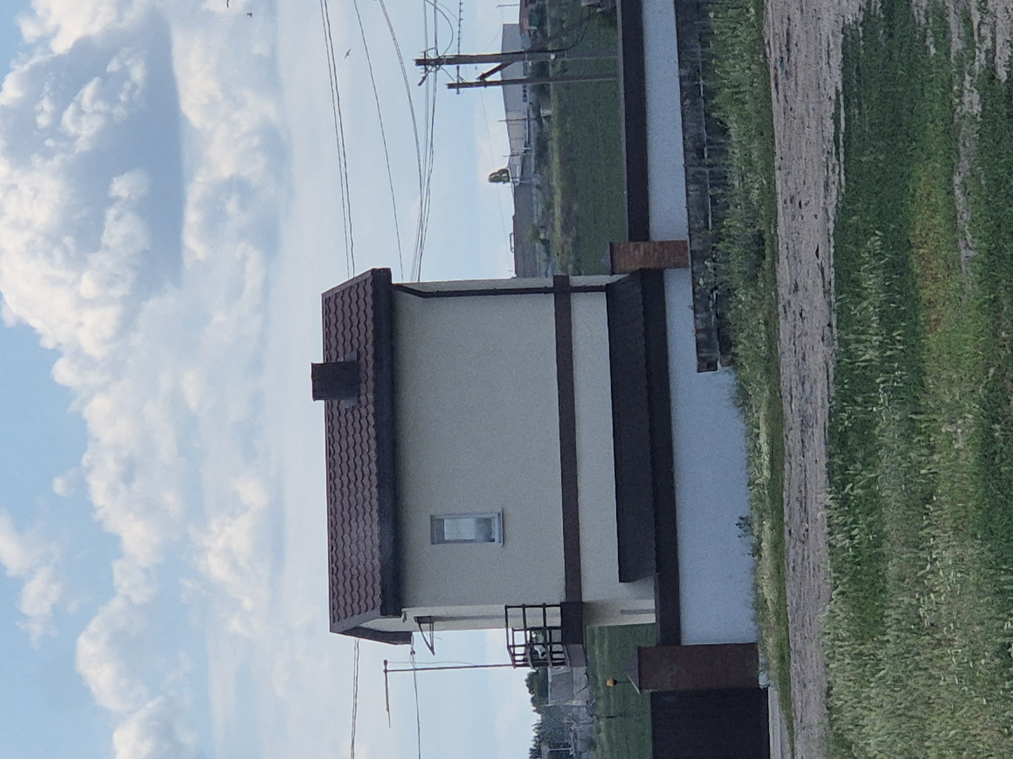The image shows a two-story building with a brown tiled roof and light beige walls. A small balcony with dark metal railings is attached to the second story. A single rectangular window is visible on the second story. The building is bordered by a white wall with a brown trim, and a dark metal gate is partially visible on the left. A brick pillar stands next to the gate. The foreground features green grass and a dirt path. In the background, there are power lines, a utility pole, and some industrial buildings. The sky is blue with scattered white clouds. The scene appears to be in an urban or suburban area, possibly on the outskirts of Pidhaitsi, Ukraine, given the context. There are no people visible in the image.