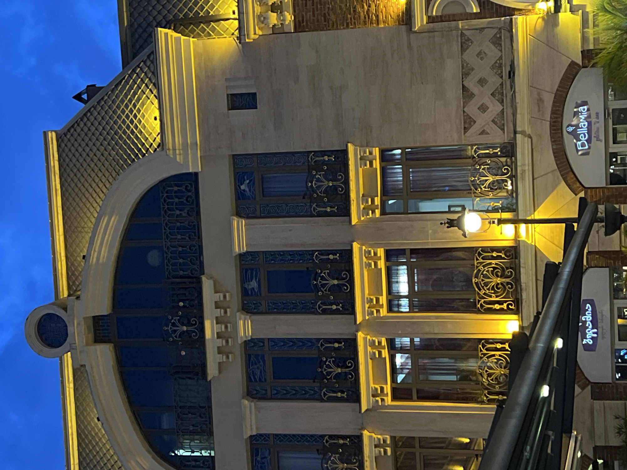 The image displays a detailed architectural facade of a building at dusk or night, brightly illuminated by warm artificial lights. The sky above is a deep, clear blue, indicating the time of day.
The building exhibits an ornate design, potentially blending Art Nouveau and classical revival styles. Its facade is constructed from a mix of light-colored stone cladding and brickwork, featuring intricate decorative patterns. A large arched window with blue panes and decorative grilles dominates the left side of the structure. To its right, several rectangular windows are visible, some revealing closed blinds or curtains inside, and all framed by richly embellished golden or brass-colored grilles adorned with swirling, organic motifs.
Additional decorative patterns are embedded within the stone work, including geometric cross-hatching and diamond shapes. Below some windows, panels with blue and white designs, resembling stylized birds or marine life, are present. On the upper left, a small, dark, triangular object protrudes from a textured, scaled roofline.
In the foreground on the right, a black lamp post with a spherical light fixture is partially visible. Two commercial signs are prominently displayed on the building's right side. The upper sign, which is curved, reads "Bellavia" with smaller text "golden Time" underneath. A rectangular sign directly below it also displays "Bellavia" in Georgian script, "ბელავი", followed by "golden Time."
No human activity is depicted; the scene is static, focusing on the architecture and its nighttime illumination. The clear sky and the specific text on the signs ("Bellavia" and "ბელავი") serve as location cues for Batumi, Georgia. The overall impression is that of an elegant and meticulously maintained urban structure, dramatically highlighted by its evening lighting.