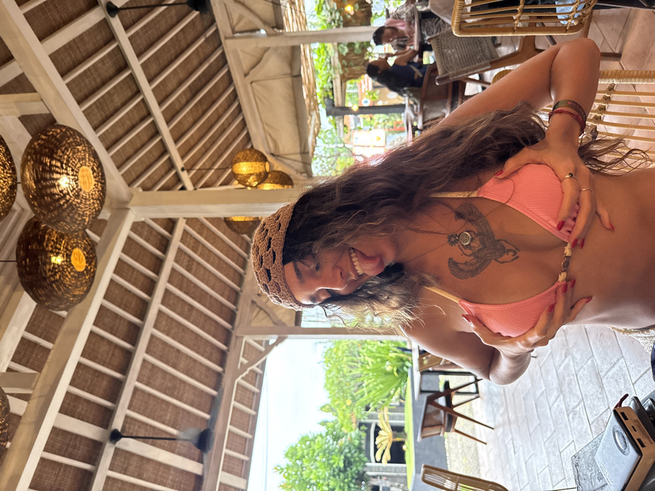 A young adult female is depicted in a semi-open-air restaurant or cafe located in Uluwatu, Indonesia. She is positioned centrally, looking towards the viewer with a playful expression, her tongue extended and a smile on her face. Her long, wavy brown hair is partially covered by a light brown crocheted cap. She wears a pink ribbed bikini top with a light-colored trim and a small central clasp. A prominent black tattoo of a winged, possibly scarab-like, figure is visible on her upper chest, beneath which she wears multiple necklaces, including one with a circular pendant. Her hands are placed under her breasts, appearing to cup or uplift them. She has multiple rings on her fingers and several colorful bracelets on her right wrist; her fingernails are painted red. She is seated at a dark-colored table, with an electronic device visible to her left.
The establishment features a high, light-colored wooden-beamed roof with woven ceiling panels and several large, spherical woven pendant lamps emitting warm light. Black ceiling fans are also visible overhead. White pillars support the roof structure. In the background, lush green tropical foliage and trees are visible, along with other patrons seated at tables. On the right side of the frame, two individuals, one wearing a dark top and another in a lighter, possibly pink, garment, are seated at a circular table. The overall environment suggests a casual, relaxed tropical dining setting illuminated by daylight.