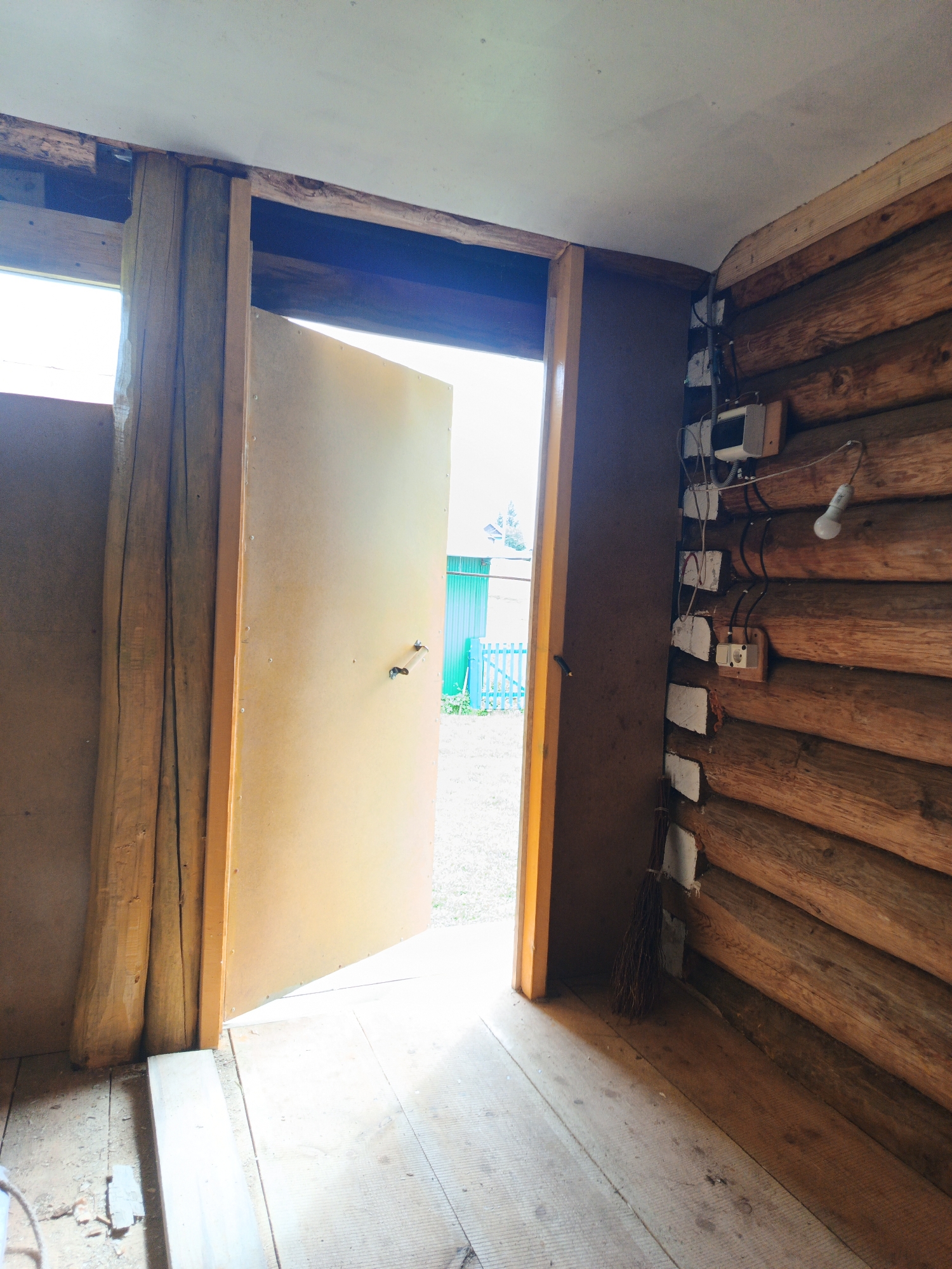 The image shows the interior of a rustic wooden structure, possibly a shed or an unfinished room, in Islambaevo, Russia. An open, light-colored door reveals a bright outdoor scene with a blue fence and green vegetation. The interior walls are constructed from rough-hewn logs. On one wall, electrical wiring and fixtures are visible, including a light bulb, an outlet, and a junction box, indicating some level of electrical installation. A broom is leaning against the log wall near the doorway. The floor is made of wooden planks, showing signs of wear and debris. The overall impression is of a functional, yet unfinished or rustic, living or working space.