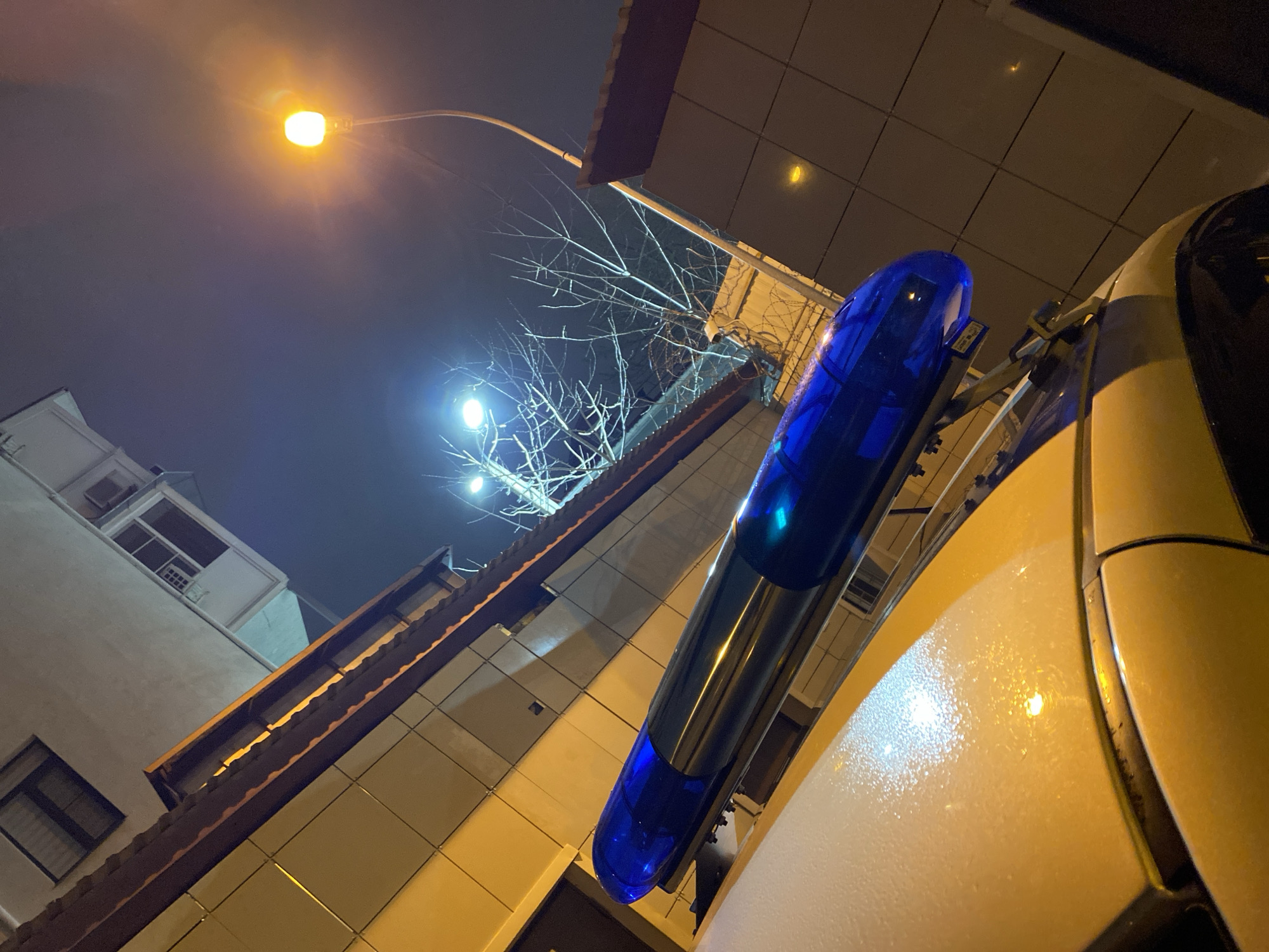 The image shows a low-angle view at night, likely in Sochi, Russia, judging by the architecture and the presence of a police vehicle with a blue light bar. The scene is illuminated by streetlights, one emitting a yellow glow and another a brighter, more diffused light. A building with a light-colored facade and a dark brown roof is visible on the left. Bare tree branches are silhouetted against the night sky. The primary subject is the police vehicle, with the blue light bar prominently displayed. The car is light-colored, possibly white or beige. The overall impression is of an urban environment under nighttime conditions.