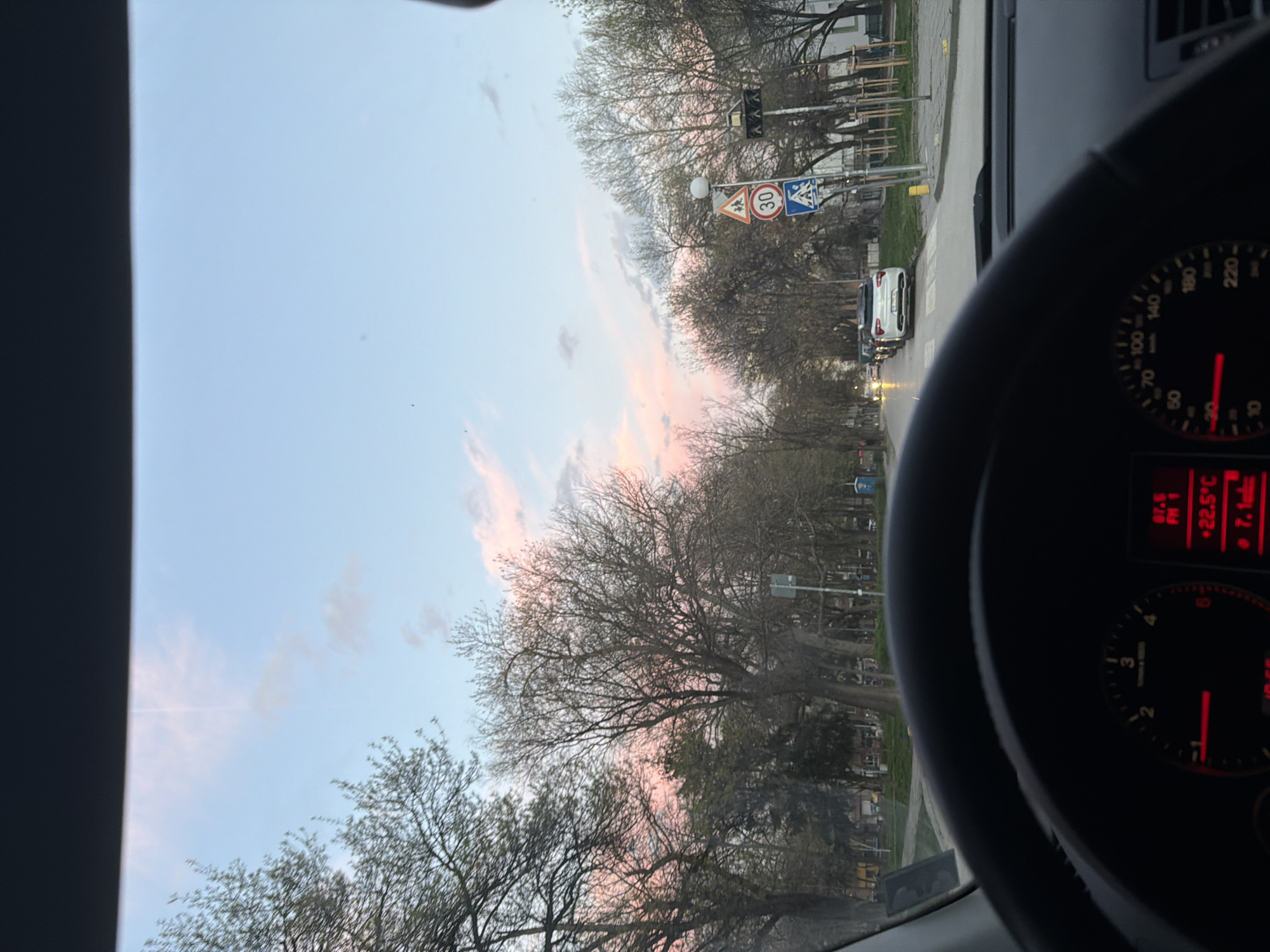 The image shows a view from inside a car driving on a street in Plovdiv, Bulgaria. The sky has a pink hue, suggesting either sunrise or sunset. Trees line the street. There are street signs indicating a speed limit of 30 km/h and a pedestrian crossing. A row of cars can be seen ahead on the road. The car's dashboard is partially visible, displaying the temperature of 22.5 degrees Celsius. A tachometer reads a low RPM, suggesting the car is either idling or moving slowly. A radio frequency of 87.6 FM is displayed.