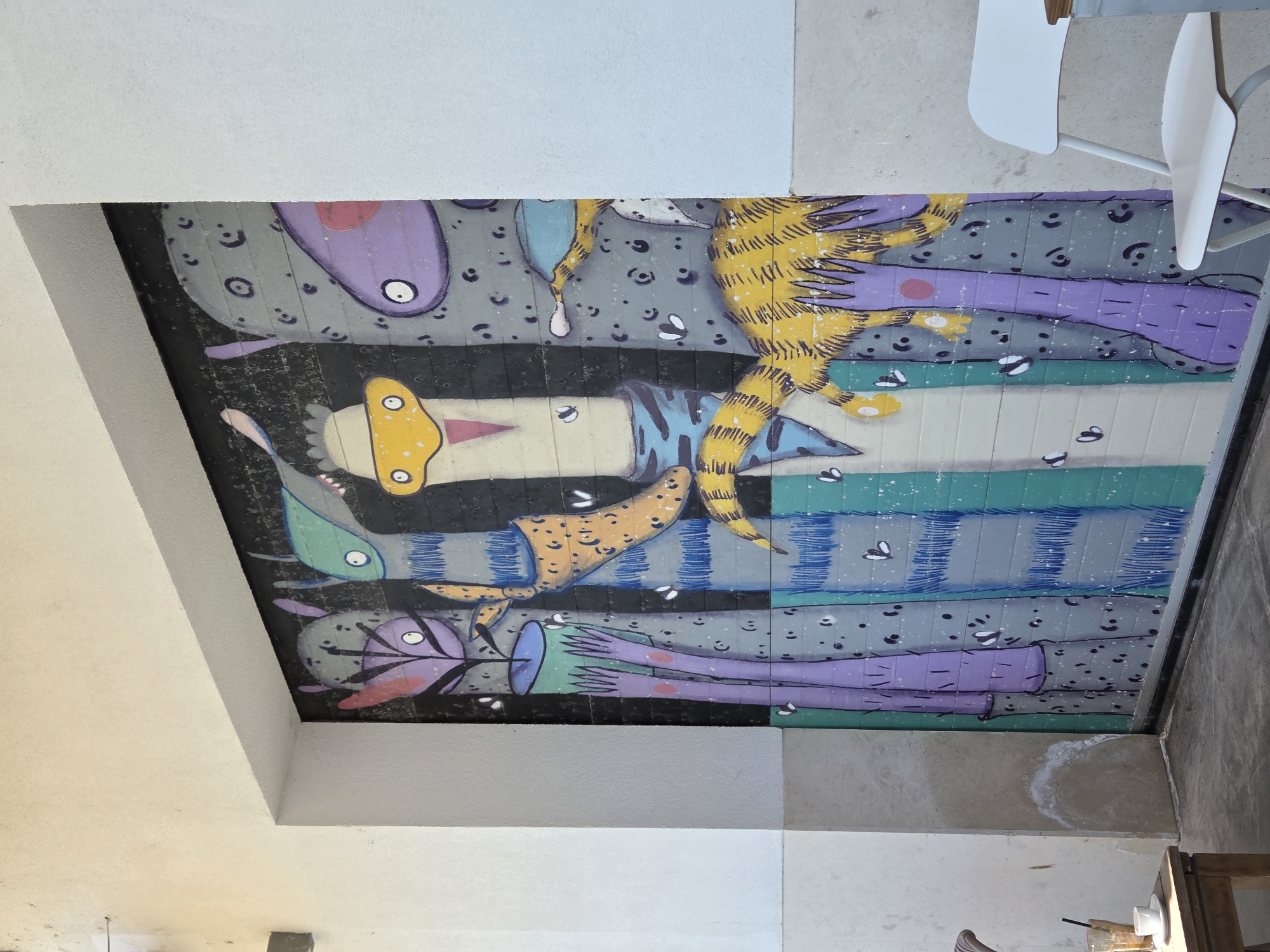 The image depicts a colorful, cartoon-style mural painted on the ceiling of a building in what is most likely Varna, Bulgaria (though this cannot be determined from the image alone). The mural features several stylized figures with elongated limbs and oversized heads. The colors used are vibrant, including shades of purple, yellow, green, and blue. The figures are arranged on a background of grey, black, and white, with small details such as dots and stripes adding to the overall design. The ceiling itself is part of an indoor space, indicated by the presence of a white wall and what appears to be the corner of a room. The lighting is bright, illuminating the mural and the surrounding area. A partially visible white chair indicates that the location may be a public space, such as a restaurant or cafe. The painting is oriented horizontally.