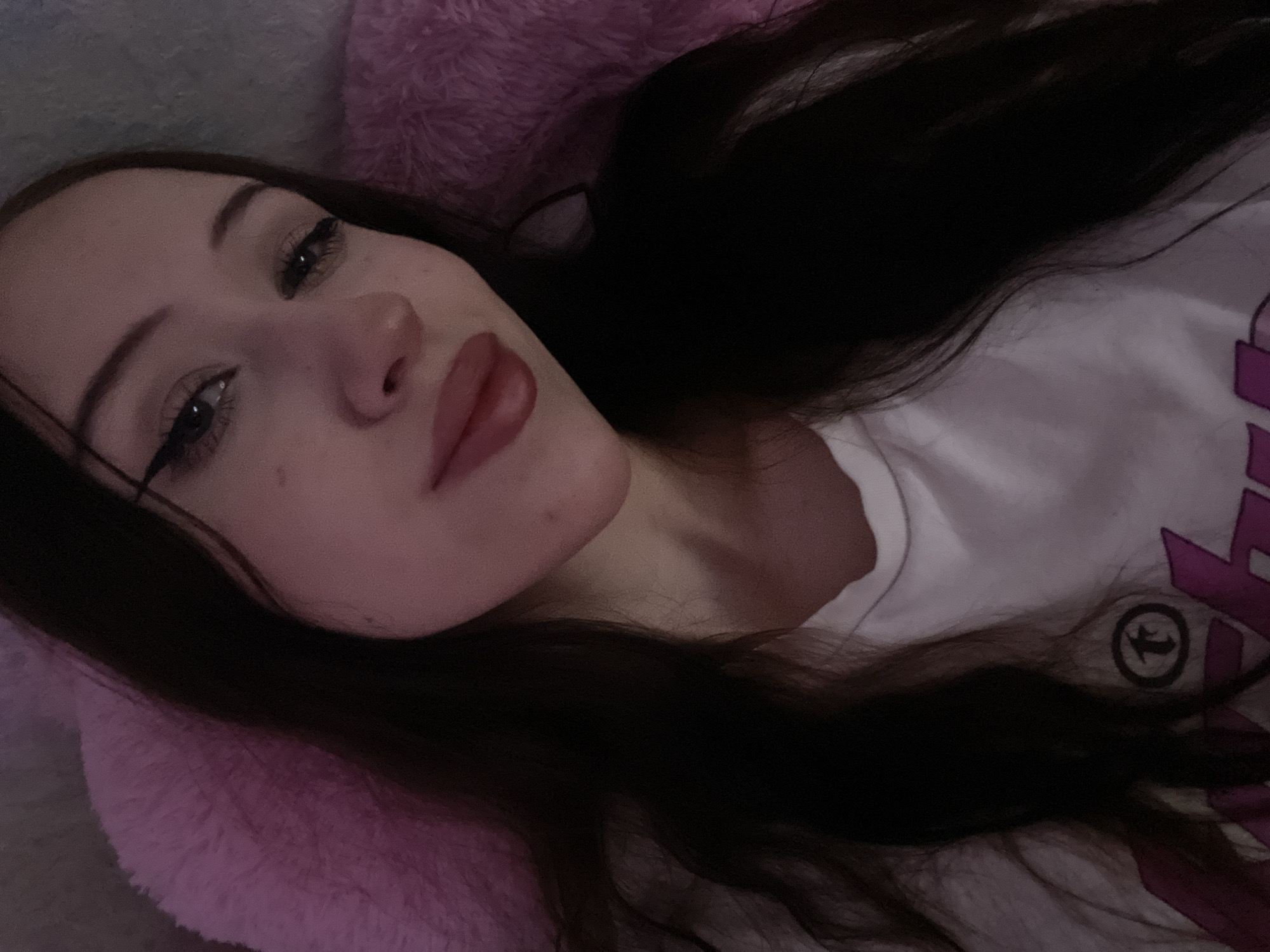 A young woman with long, dark hair is lying down, resting her head on a fuzzy pink pillow. She is wearing a white t-shirt with a graphic print. Her makeup includes eyeliner and lipstick. The lighting is dim, suggesting an indoor setting, likely a bedroom. The image is a selfie, taken from a slightly upward angle. There is no information in the image to indicate the location in Votkinsk, Russia.