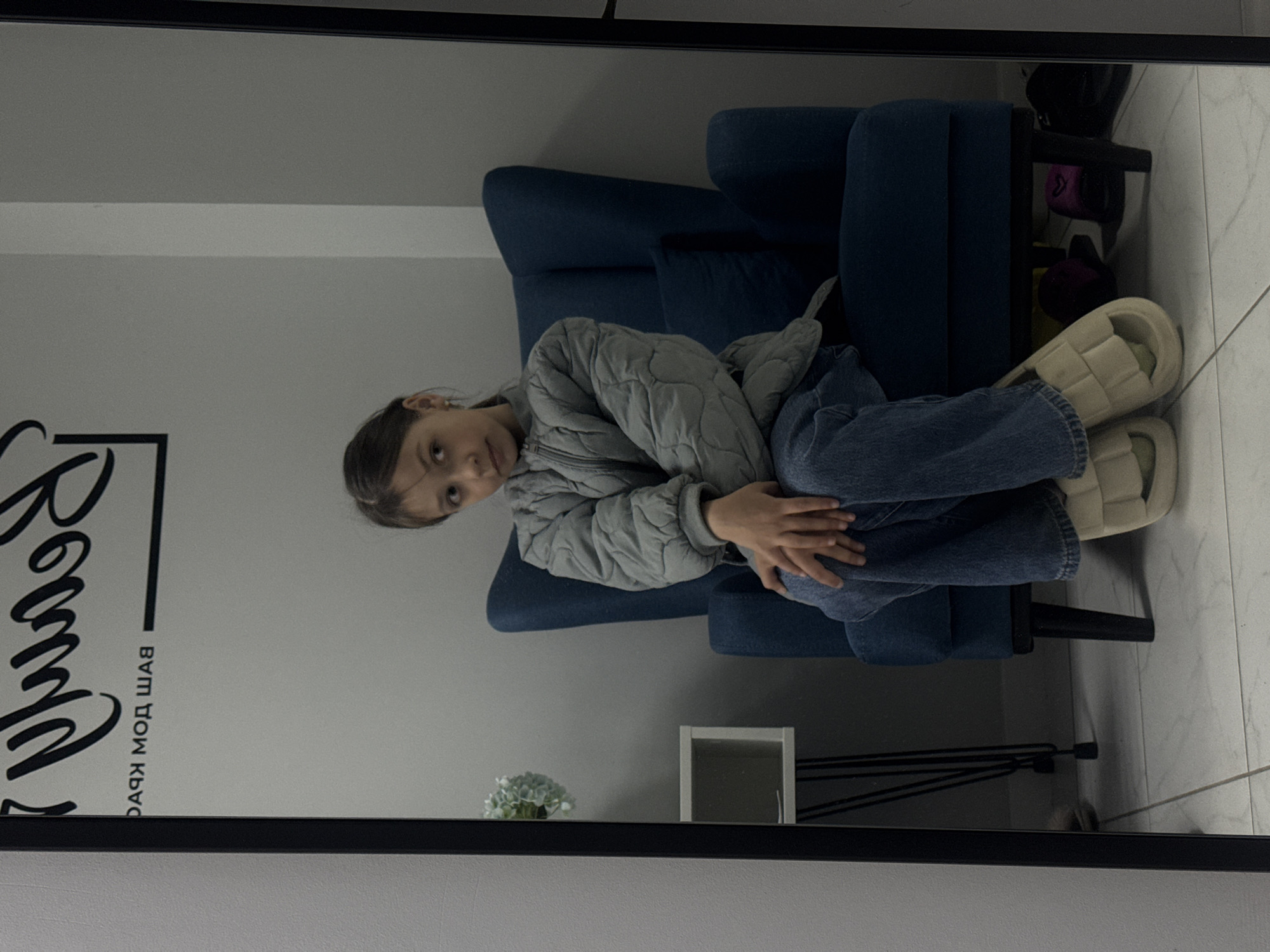 A young girl wearing a quilted grey jacket and blue jeans is seated on a blue armchair. She is reflected in a mirror. The reflection also includes part of a wall with a logo or sign written in what appears to be Russian. A small flower arrangement and a small white square decorative item are visible below. To the right of the armchair in the reflection, there is a black side table with items on the shelves, including footwear, and a pair of off-white quilted slippers are on the girl's feet. The floor appears to be white tile. The mirror has a black frame. The location appears to be an indoor setting in Votkinsk, Russia, possibly a beauty salon or similar establishment.
