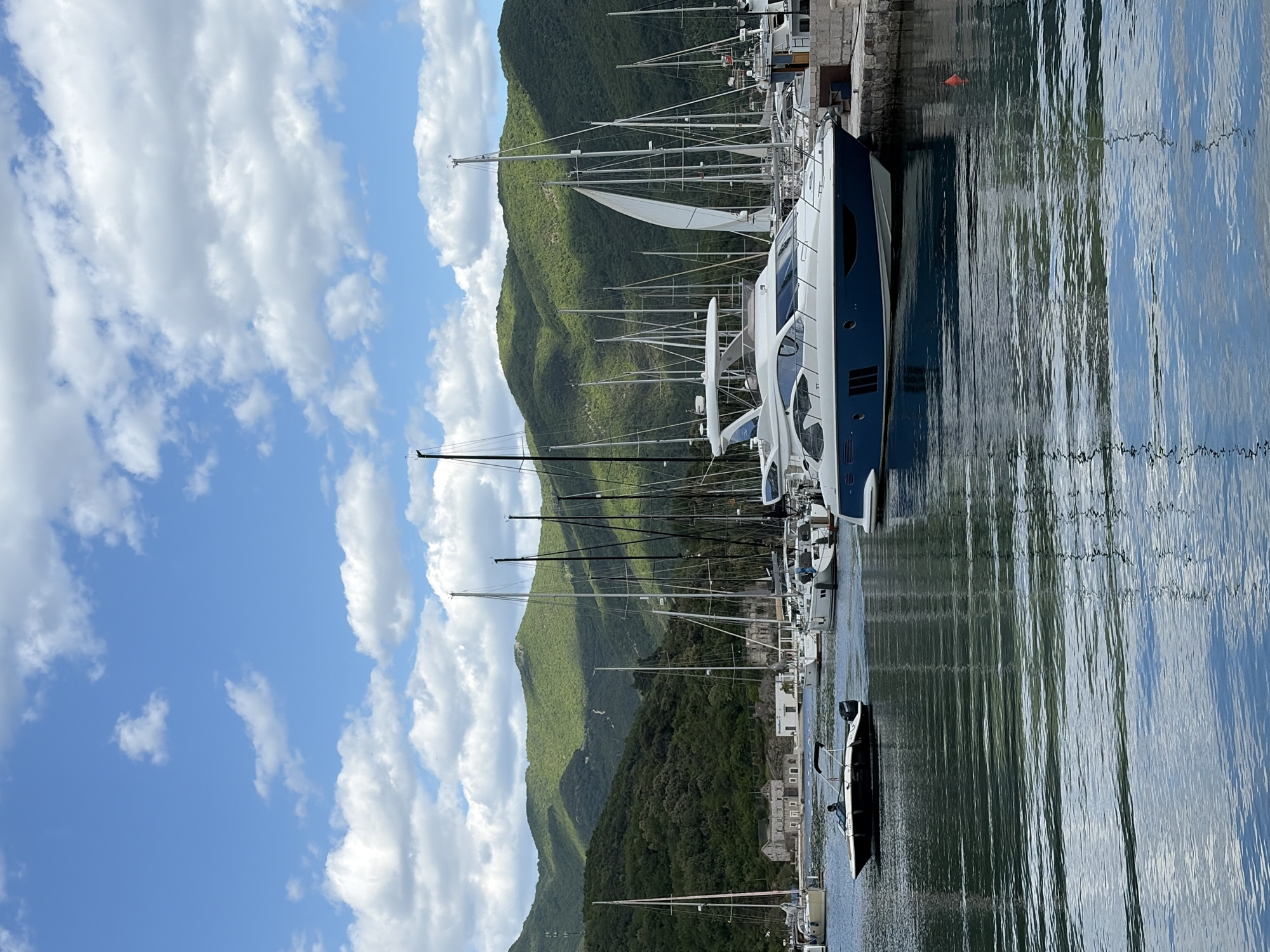 The image shows a harbor scene in Herceg Novi, Montenegro. Several sailboats and yachts are moored at a pier along the harbor. A backdrop of mountains covered in green vegetation rises behind the harbor and pier. The sky is blue with scattered white clouds. The water in the harbor has reflections of the sky and boats.
