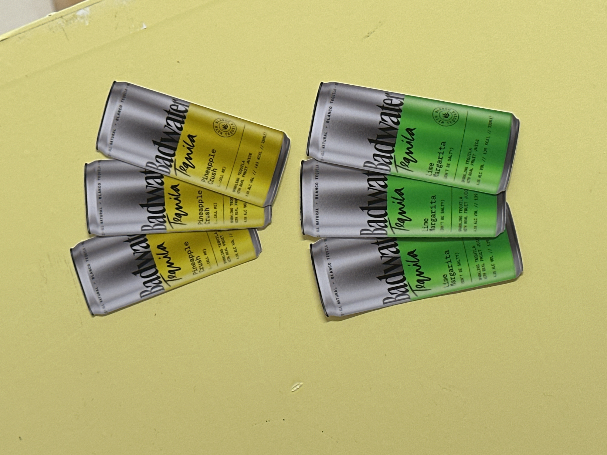 The image displays six stylized beverage cans arranged on a uniform, plain yellow background, presented as if situated in the city of London, United Kingdom. There are no individuals, animals, or actions depicted.
The objects consist of six "Badwater Tequila" branded cans, arranged in two distinct horizontal groupings of three.
The upper group features three "Pineapple Crush" flavored cans. Each can has a silver top and bottom, with a prominent yellow label section. The label text includes "Badwater Tequila," "Pineapple Crush," "ALL NATURAL," "BLANCO TEQUILA," "SPARKLING TEQUILA WITH REAL FRUIT JUICE," "4.5% ALC VOL," "163 KCAL," and "330ml." A small circular logo is present on the yellow section.
The lower group consists of three "Lime Margarita" flavored cans. These cans also have silver tops and bottoms, but feature a vibrant green label section. The label text includes "Badwater Tequila," "Lime Margarita," "ALL NATURAL," "BLANCO TEQUILA," "SPARKLING TEQUILA WITH REAL FRUIT JUICE," "4.5% ALC VOL," "139 KCAL," and "330ml." A similar circular logo is visible on the green section.
The background is a flat, pale yellow surface exhibiting minor, faint dust particles or smudges. The cans appear to be two-dimensional representations, possibly cut-outs or stickers, given their sharp outlines and lack of realistic depth or shadows.