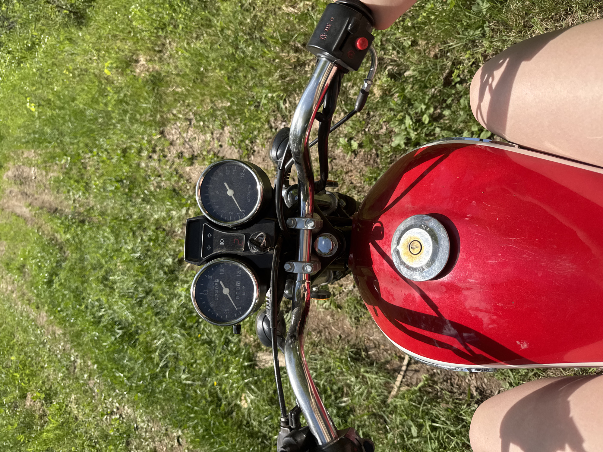 The image depicts the view from the handlebars of a red motorcycle, likely in a grassy, outdoor setting. The handlebars are chrome and feature standard motorcycle controls, including switches and a throttle. In front of the handlebars are two gauges, one displaying speed in km/h and the other displaying RPM. The motorcycle's red fuel tank is prominent in the foreground. A person is sitting on the motorcycle; only their thighs are visible in the frame. The environment is a grassy area, possibly a field or park. There's no direct evidence to confirm the location as Zhigalki, Russia, from the given image content.
