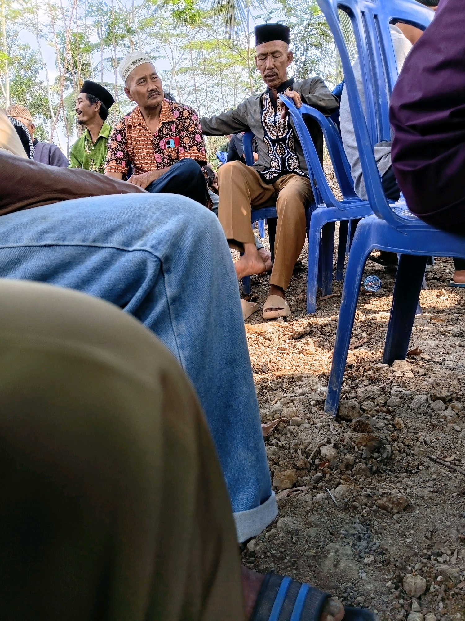 A group of men are seated outdoors in Margo Rukun, Indonesia, on a dirt ground covered with stones and dry leaves. The men are wearing traditional Islamic attire, including peci caps and sarongs. They are seated on blue plastic chairs, and some are looking away from the camera. The scene appears to be a gathering or a meeting.