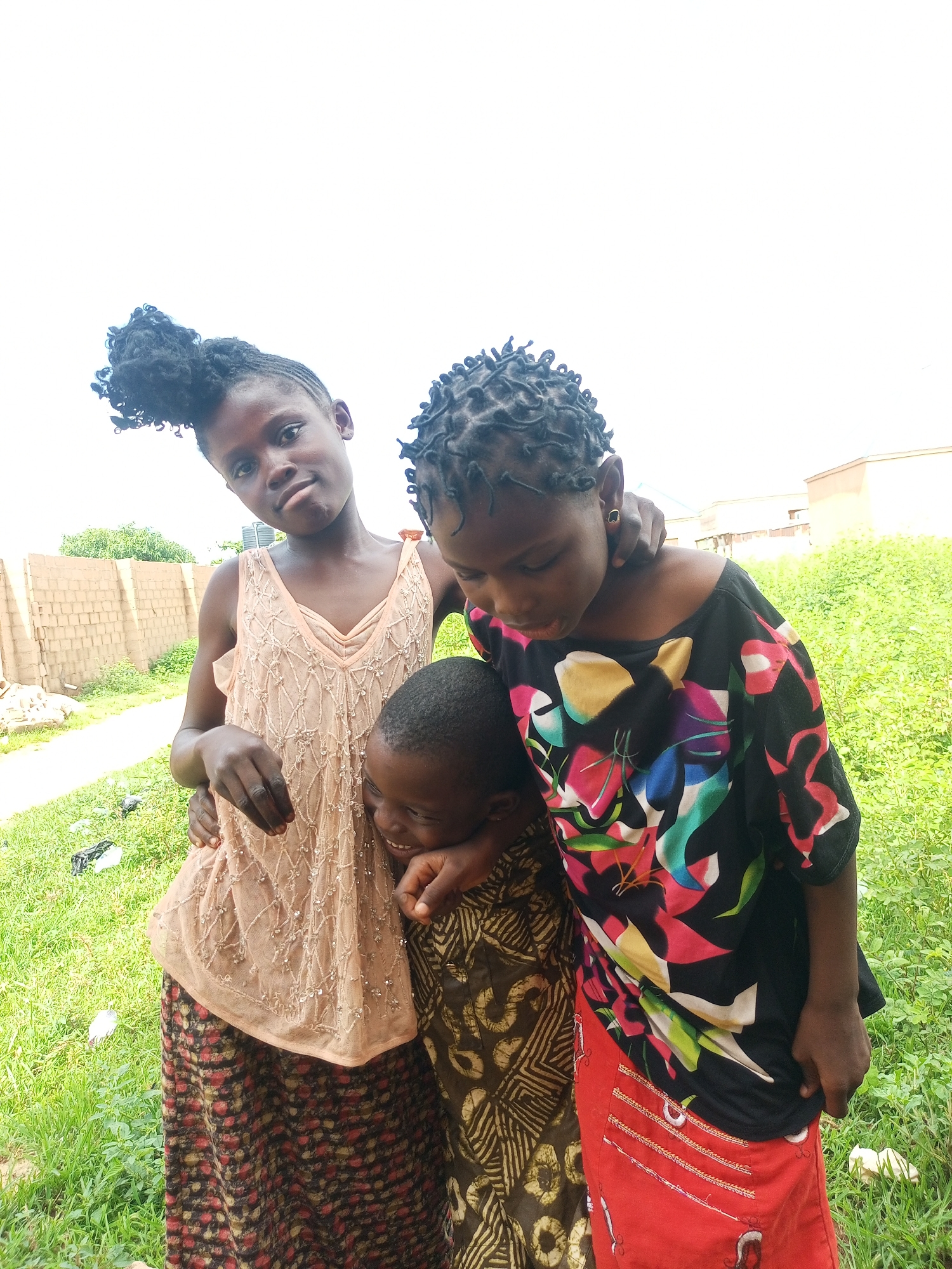 Three young Black children are posed together outdoors, likely in Tempure, Nigeria. The child on the left, a girl, has her dark hair styled in a high bun with curly tendrils. She wears a light peach-colored, sleeveless dress with a V-neck and a beaded pattern, and a long, patterned skirt. Her left arm is around the neck of the child in the center. The child in the center is younger and appears to be a boy, with short, dark hair. He is wearing a brown and tan patterned shirt and is leaning into the child on the left, smiling. The child on the right is a girl with dark hair styled in small braids or twists. She is wearing a black t-shirt with a vibrant, abstract floral pattern in pink, yellow, green, and blue. Her right arm is around the neck of the child on the left. She is looking down and smiling.
The background consists of a low, tan brick wall on the left, with some greenery behind it. Further in the background, there is a body of green grass and some sparse trees. The sky above is overcast and bright white. The ground is covered with grass, with some scattered litter visible. The lighting suggests it is daytime, possibly morning or late afternoon, with the sun being diffused by clouds. The overall impression is of a casual, outdoor gathering.