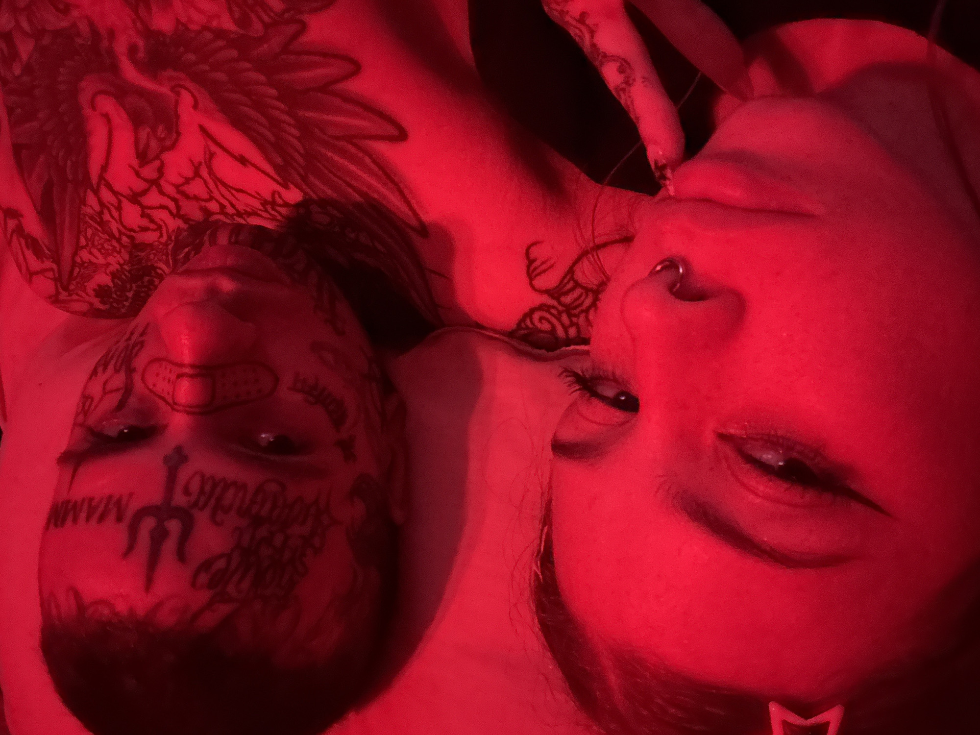 Two people are lying down next to each other, illuminated by red lighting. The person on the left has multiple face tattoos, including what appears to be the word "Miami" and a trident. The person on the right has a nose ring and a hand with tattooed fingers near their face. The location is unclear and cannot be determined to be Buenos Aires, Argentina, based on the given image.