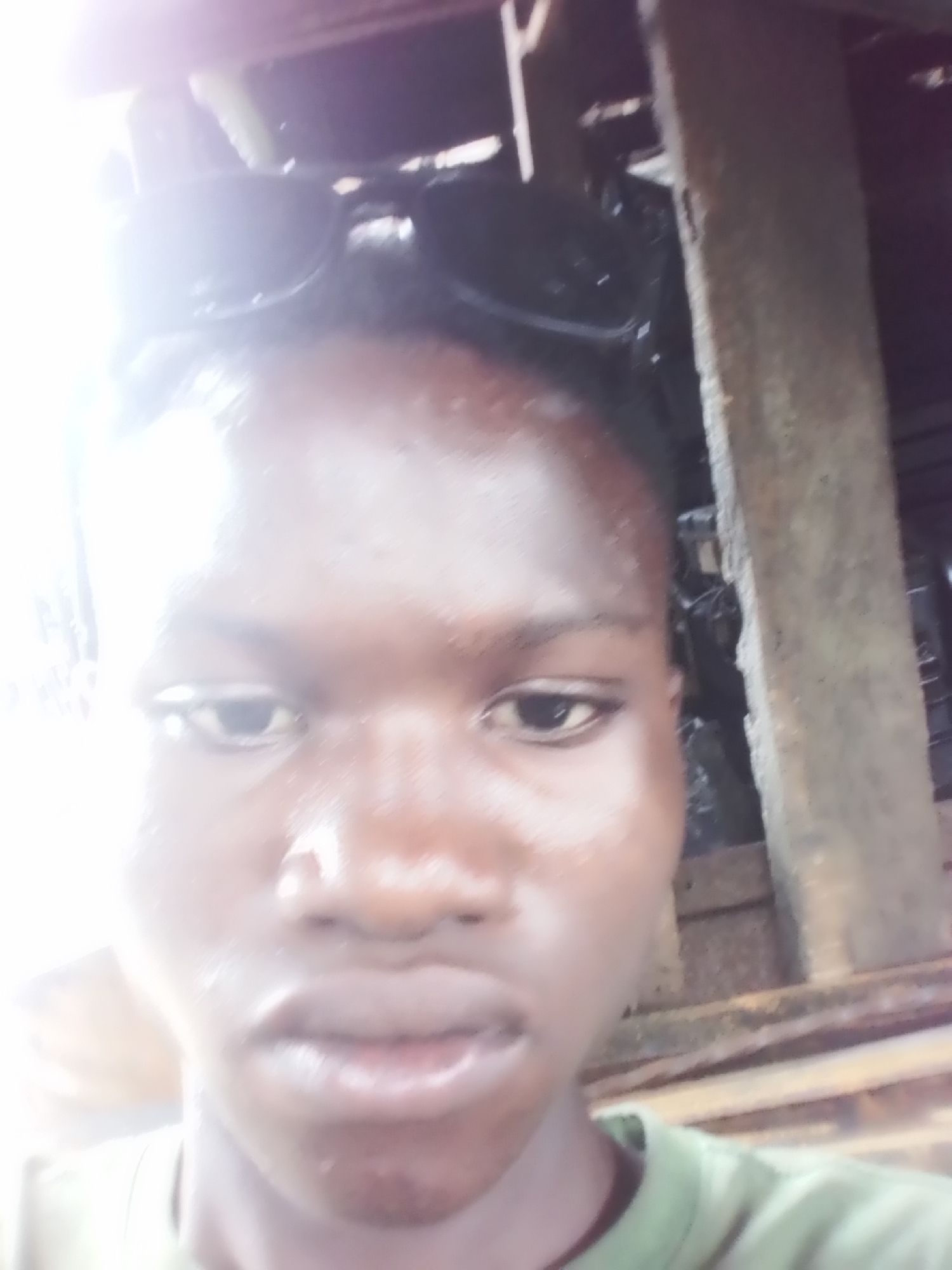 The image is a close-up, slightly blurred selfie of a person, likely a young male, with dark skin, looking directly at the camera. He is shown from the upper chest upwards. A pair of dark sunglasses rests on top of his head. His face appears shiny, possibly from sweat or the intense lighting. The expression on his face is neutral.
The left side of the image is severely overexposed with a bright, white glare that washes out details, particularly on the left side of his face and the background. In contrast, the right side of the background is darker and shows rough, dark wooden planks or beams, suggesting a rustic or industrial setting, possibly indoors or in a sheltered outdoor area with wooden construction. No specific activity is depicted other than posing for the photo. There is no visible text in the image.