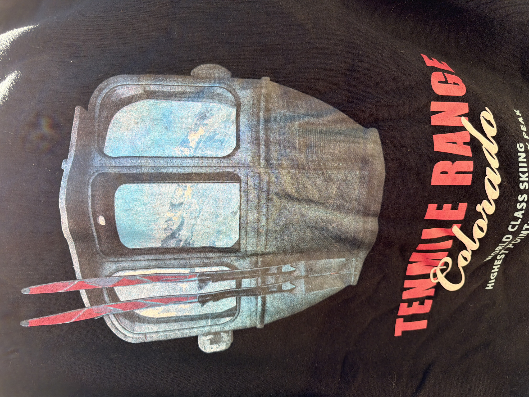 The image shows a black T-shirt with a print on it. The print features a gray and blue gondola-like structure with skiis attached. To the right of this image, in pink, are the words "Tennille Range Colorado". Below that is written "WORLD CLASS SKIING PEAK HIGHEST POINT".