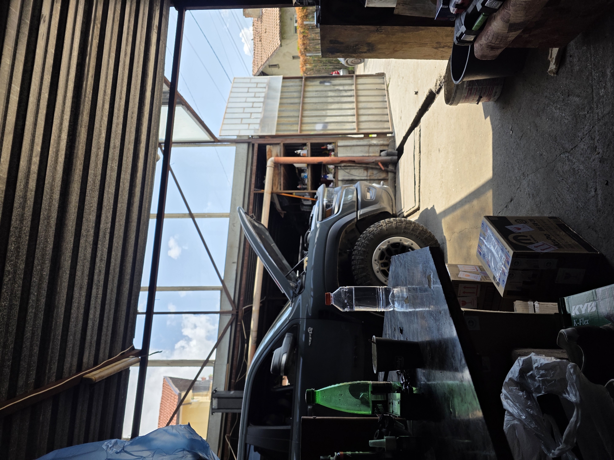 The image captures an interior view of a workshop or garage, looking out towards an outdoor area under bright daylight in Dragichevo, Bulgaria.
In the foreground, a dark workbench or counter occupies the right side, laden with various items. These include a clear plastic water bottle, a green plastic bottle with partial text "HEMAN," and what appears to be a clear glass or cup. Stacked cardboard boxes are present, with visible branding such as "MOPAR" and "KYB K-Flex," suggesting car parts or accessories. A white plastic bag is also seen on the far right.
The central focus of the midground is a dark-colored vehicle, possibly an SUV or truck, with its hood propped open, indicating maintenance or repair work. Its robust, off-road-style tires are visible on the right side of the vehicle.
To the left, the internal structure features corrugated metal siding or roofing. The workshop space is brightly lit by natural light streaming in from a large opening, revealing a clear blue sky with some clouds overhead. Outside the workshop, a building with a reddish-tiled roof can be seen in the distance, alongside another structure composed of white bricks and corrugated metal panels. Power lines are faintly visible against the sky.
No individuals are present in the scene. The overall impression is that of an active, functional vehicle repair or maintenance facility on a sunny day.