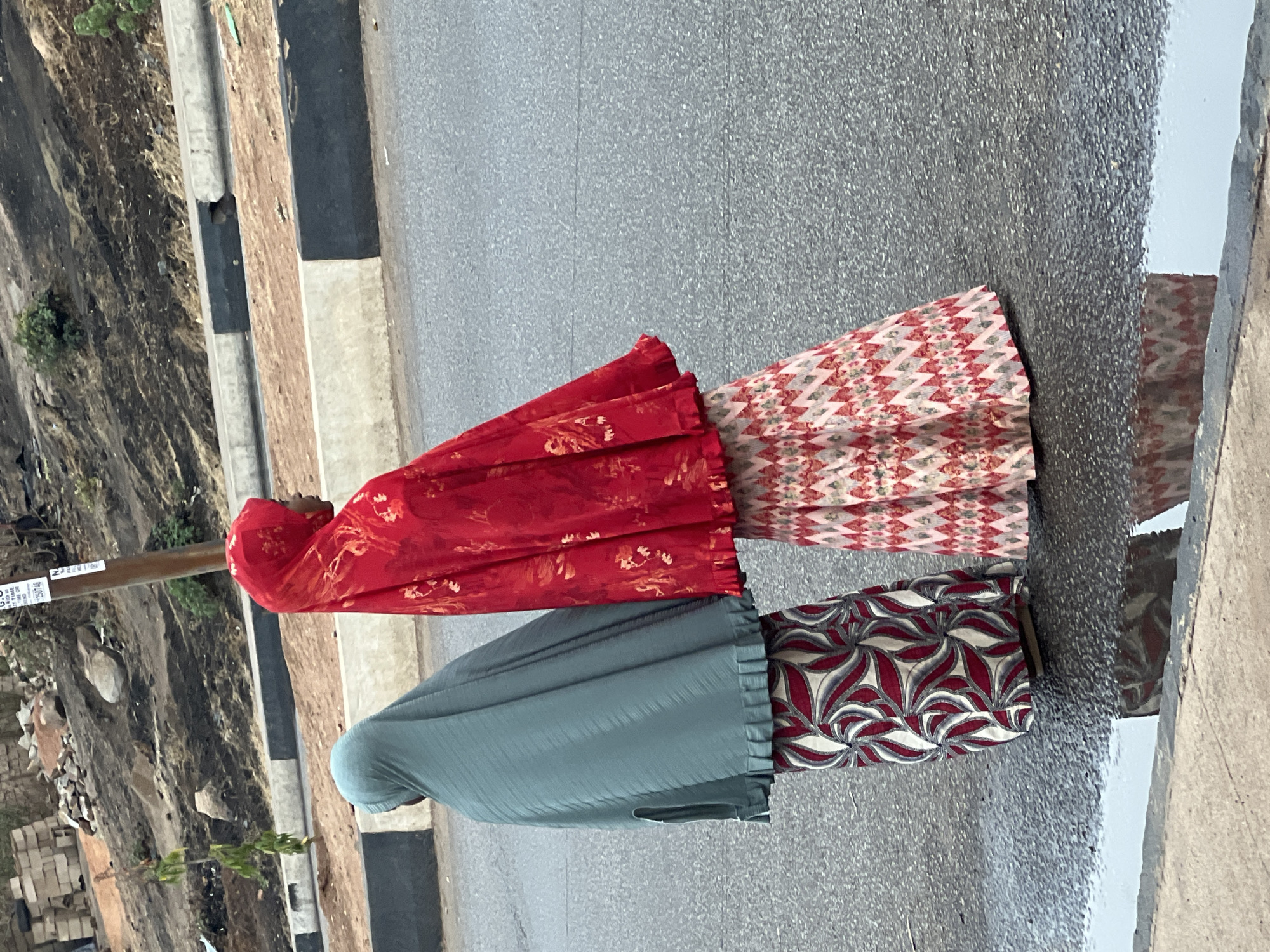 Two women stand next to a road in Jos, Nigeria. One woman is wearing a red head covering and a patterned skirt. The second woman is wearing a gray head covering and a patterned skirt. The road appears wet. There is a vegetated, excavated area beside the road.
