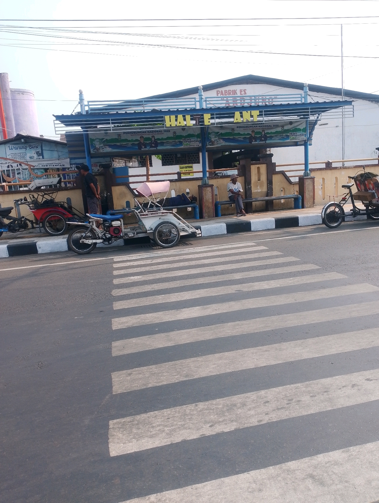 The image captures a street scene in Probolinggo, Indonesia, featuring a bus stop with a prominent sign that reads "HALTE ANT" and "PABRIK ES ARDJUNO." Multiple motorized tricycles, commonly known as "becak motor," are parked near the bus stop. One becak motor is white with a blue seat and a red engine visible. Another is red with a similar structure. A third, a bicycle with a cargo area at the back, is parked to the right.
Two individuals are seated on a bench at the bus stop. One is a man wearing a hat, a white shirt, and dark pants, looking down. Another person, partially visible, is lying down on the bench. A man in a dark t-shirt and shorts stands near the entrance of the bus stop structure.
A pedestrian crossing with alternating white and gray stripes is prominent in the foreground, indicating a road. The environment appears to be outdoors with a clear sky and buildings in the background. A sign near the bus stop entrance has text in Indonesian, including "SELAMAT DATANG" (Welcome) and "BARANG MASUK" (Goods Entry).