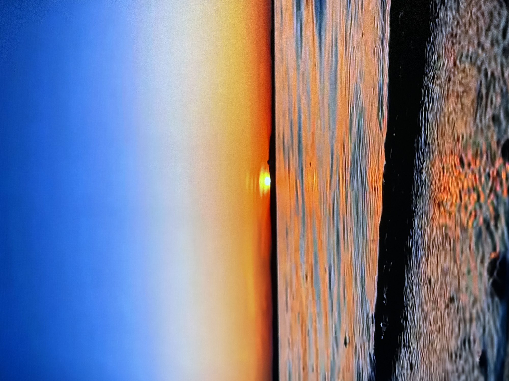 A seascape at dusk features a vibrant sunset over a calm body of water. The sky transitions from a deep blue at the top to shades of orange and yellow near the horizon. The sun, a bright yellow orb, is partially visible, casting a golden reflection on the water's surface. The water itself is a mix of blues and oranges, with gentle ripples creating a textured effect. A dark, curved line, possibly a wave or a shadow, runs along the bottom of the frame, separating the water from the foreground where small, dark objects are scattered. The overall scene is serene and visually striking, evoking a sense of tranquility.