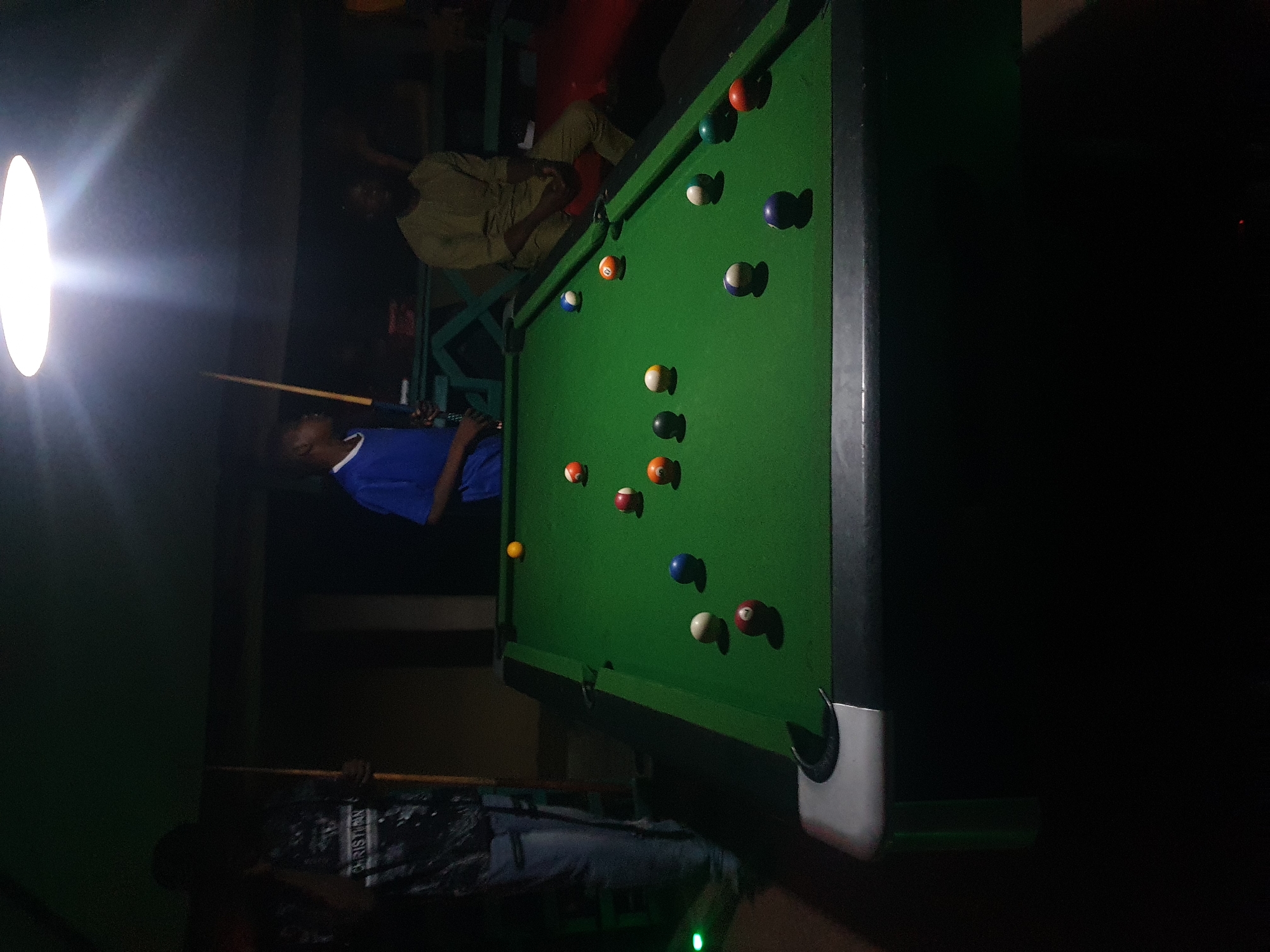 The image shows a pool table in what appears to be an indoor setting, likely a game room or bar, with dim lighting. The pool table is green and has several billiard balls scattered across it, including solid colors and striped balls. Some balls are numbered.
In the foreground, a person wearing a blue shirt is leaning over the pool table, holding a pool cue, and appears to be playing. Their face is not clearly visible. Further back, another person is seated or leaning near the pool table, wearing a light-colored shirt. The background is dark and indistinct, but suggests an indoor environment with some structural elements and possibly other people further away.
The overall mood seems relaxed and casual, typical of a game of pool. The lighting suggests it might be evening or nighttime. There are no obvious indicators of the weather. The location context of Lokoja, Nigeria is provided, but no specific visual cues in the image strongly suggest this particular location.
One of the cropped images reveals a portion of text that appears to say "CHRISTIAN" on a dark patterned garment in the lower part of the image, worn by a person whose face is not visible.