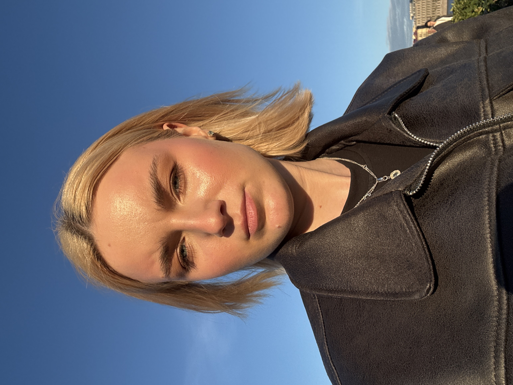 This media file features a close-up, rotated portrait of a young woman with blonde hair against a clear blue sky. She is wearing a dark, textured jacket, possibly leather or faux leather, with a zipper and a flap pocket visible. A silver necklace with a pendant is also visible. The woman's face is turned towards the camera, and her expression appears neutral or slightly contemplative. The lighting suggests it is daytime, likely late afternoon or early evening due to the warm glow on her skin. In the background, a blurry hint of buildings and greenery suggests an outdoor urban or semi-urban setting, potentially in Горбунки, Russia. The image's orientation and the clear sky indicate an outdoor activity, possibly a selfie taken on a bright, sunny day. There is no visible text.