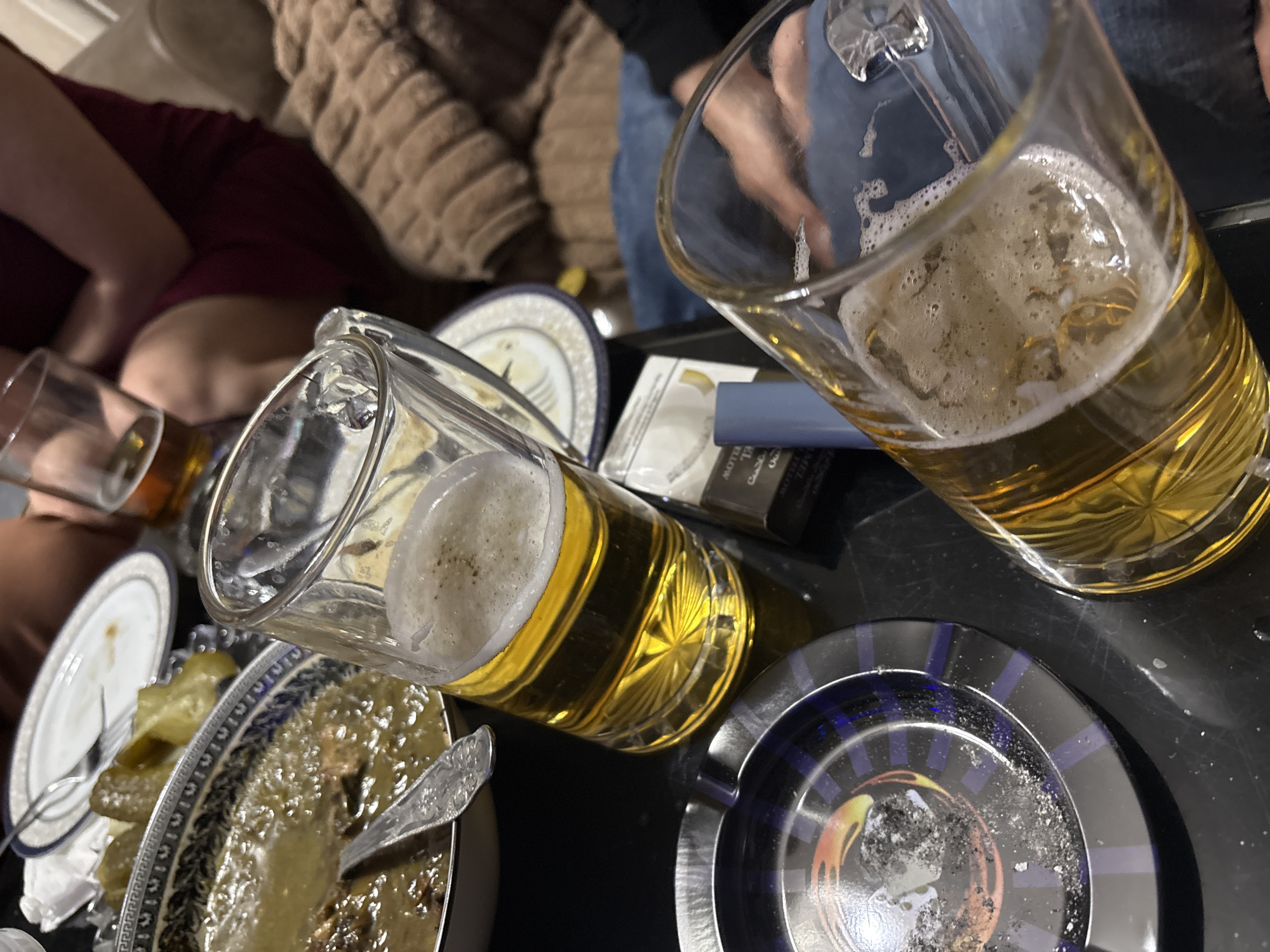 This image captures a casual, intimate indoor gathering, likely in the evening. The scene is viewed from a slightly elevated perspective, looking down at a dark table or tray laden with items.
The main subjects are two clear glass beer mugs, prominent in the foreground. The mug on the right is almost full with golden lager and a substantial foamy head, while the one on the left is slightly less full. Both mugs have a textured, patterned base.
Surrounding the beverages are several items indicating a social event. A dark blue and black ashtray with an orange and yellow circular design is visible on the right, filled with ash and discarded cigarette butts. Adjacent to the left beer mug is a white and blue cigarette packet, partially revealing the text "MOTTE" and some Arabic script.
Several dishes are also present. A decorative plate with intricate blue and white patterns holds what appears to be a hearty meal, possibly a stew, with a spoon resting within it. To its left, a small dish contains green pickles. Another white plate with a blue rim and a fork is partially visible further left.
In the blurred background, parts of at least two individuals are discernible. One person on the left is wearing a maroon garment, while another on the right is seen wearing blue jeans. A soft, brown textured blanket or cushion provides a cozy backdrop. The overall impression is one of relaxed camaraderie, focused on shared drinks, food, and conversation.