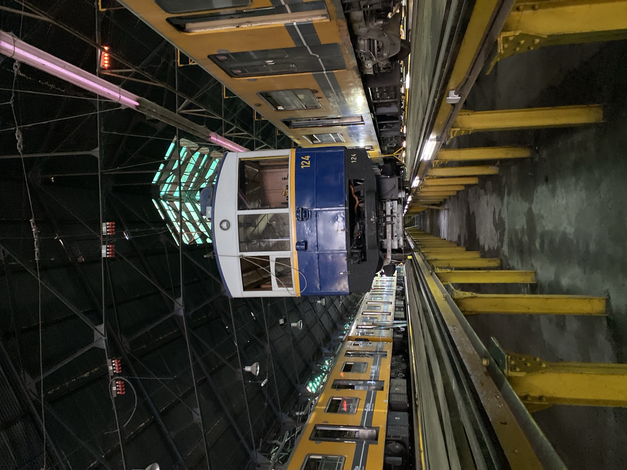 The image depicts a train maintenance facility, possibly in Buenos Aires, Argentina. Two train cars are visible, one predominantly blue and the other yellow and grey. The blue train car has the number "124" prominently displayed. The trains are positioned parallel to one another, underneath an overhead structure of metal supports, lights, and electrical components. A series of yellow structural beams runs along the right side of the frame. The floor is a dark grey, likely concrete, and appears somewhat reflective. The environment is dimly lit. The image contains no people.