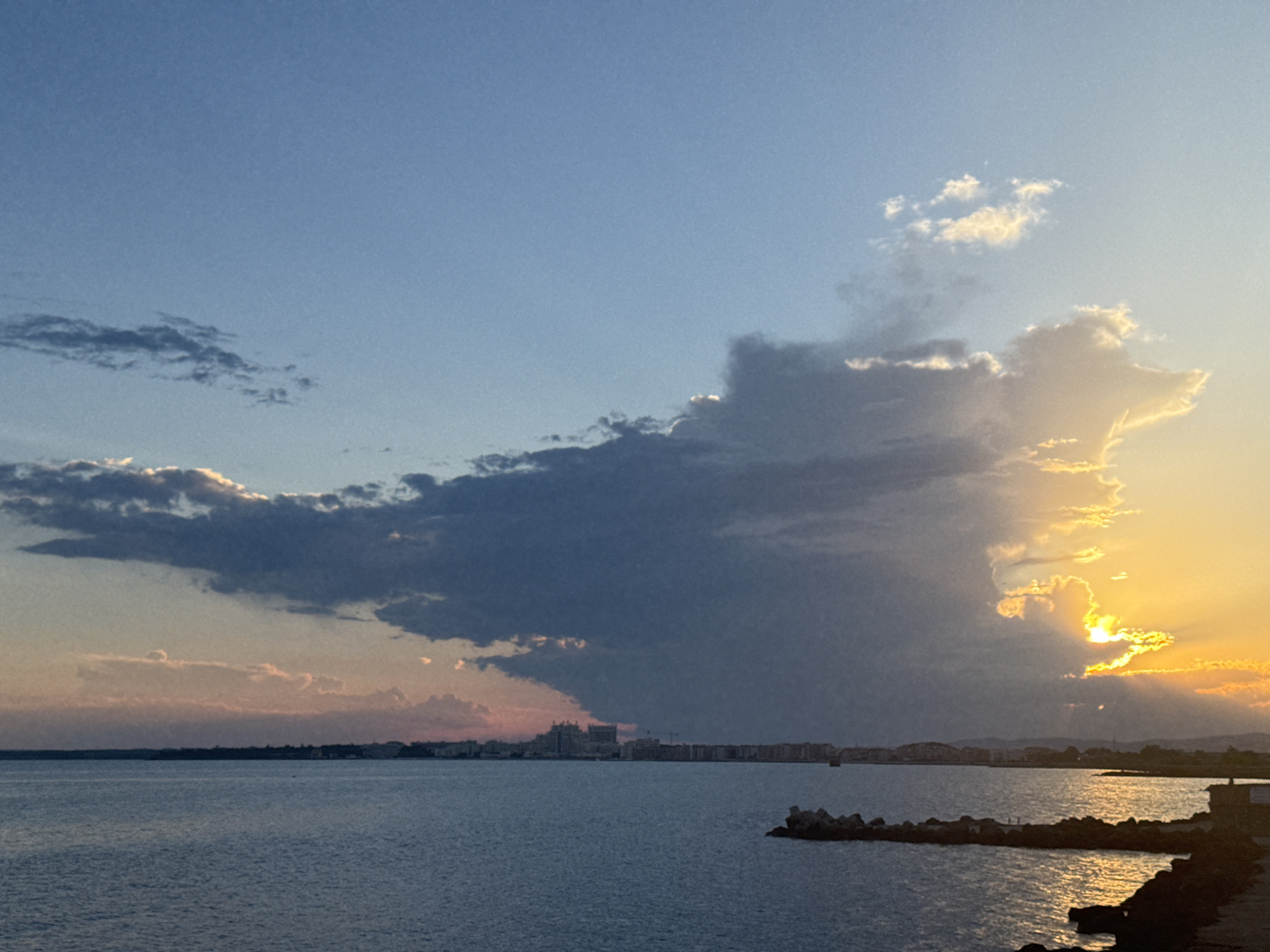 The image shows a seascape at sunset, with the sun partially obscured by clouds on the right side of the frame. The sky transitions from a clear blue at the top to warmer tones of orange and yellow near the horizon. A line of buildings, likely part of the city of Pomorie, Bulgaria, is visible along the distant coastline. The water in the foreground is calm and reflects the colors of the sky. There are some breakwater structures made of concrete tetrapods and rocks along the shoreline. No people or specific actions are discernible in the image.