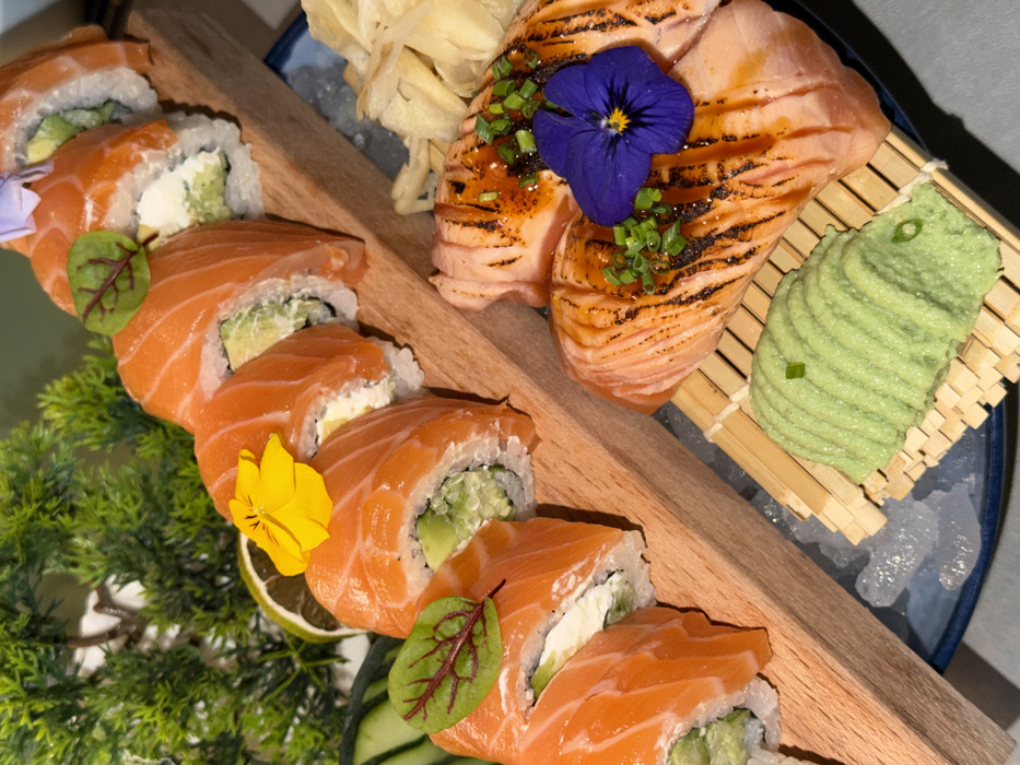 The image displays an overhead view of a sushi and sashimi platter presented on ice. The primary subjects are two distinct preparations of salmon.
On the left, a row of eight uramaki sushi rolls is arranged on a light-colored rectangular wooden board. Each roll features a white rice exterior, an inner filling appearing to include avocado and a cream-colored substance (likely cream cheese), and is topped with a thin slice of raw orange salmon. Individual rolls are garnished with small yellow edible flowers and small red-veined microgreen leaves. Sliced cucumber and a wedge of lime are visible to the left of the wooden board, accompanying a dense green, faux or live decorative plant in the blurred background.
On the right, several slices of seared or grilled salmon, exhibiting grill marks and a glossy dark sauce, are presented on a bamboo mat. These are garnished with chopped green onions and a single purple edible flower. Adjacent to the seared salmon, a mound of light green wasabi paste and several thin, pale yellow slices of pickled ginger (gari) are visible.
Both the wooden board and the bamboo mat rest on a bed of crushed ice, contained within a dark blue or black ceramic plate or bowl. The immediate background is out of focus, showing shades of green from the decorative plant and soft grey tones. No people are visible, and no discernible actions or interactions are taking place. The specific location within Budva, Montenegro, cannot be determined from the visual information provided.