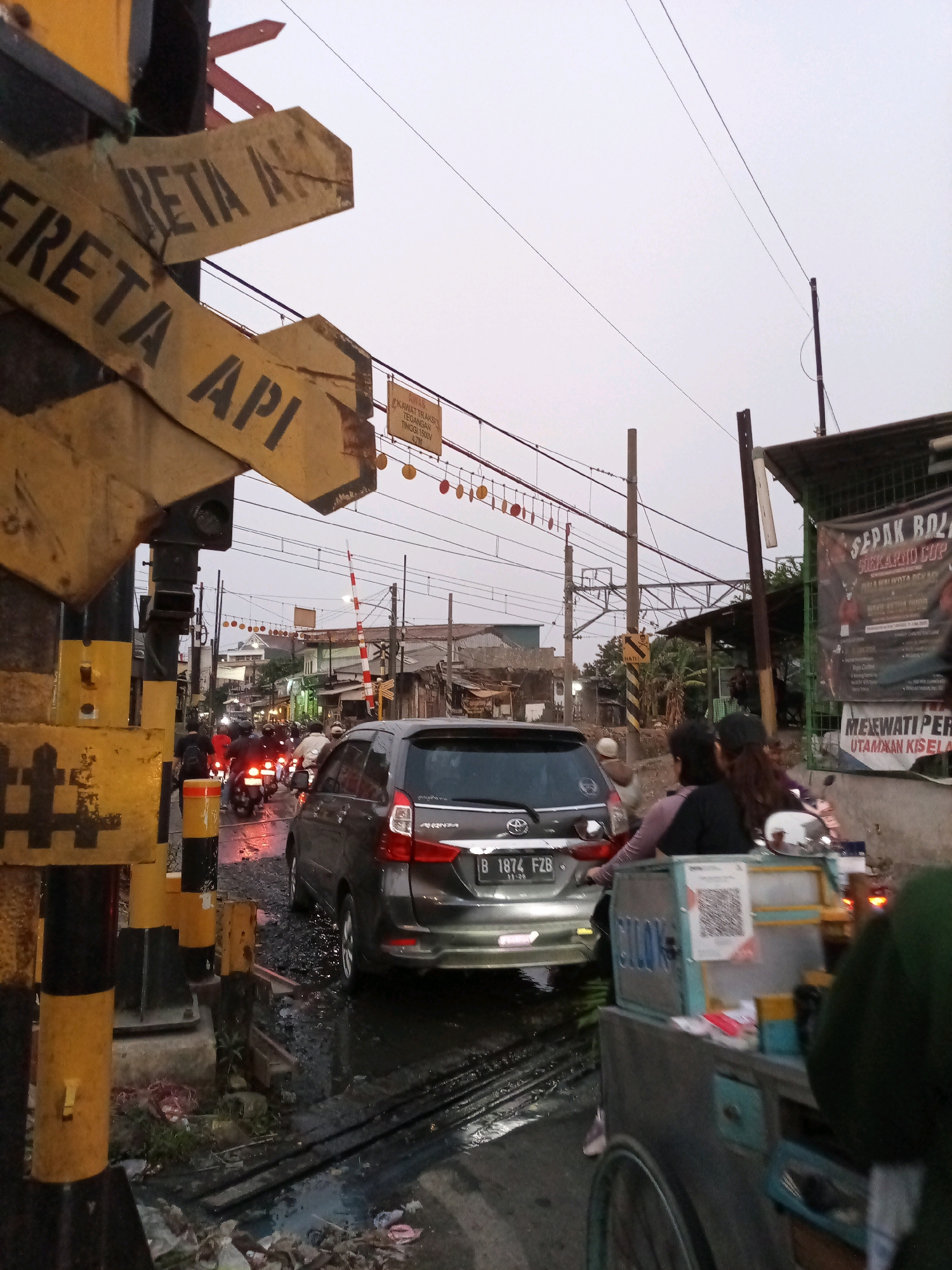 The image depicts a busy street scene at a railway crossing in Bekasi, Indonesia. In the foreground, a yellow and black striped pole, part of a railway crossing barrier system, stands prominently. Several weathered yellow directional signs with black lettering are attached to the pole, indicating "FRETA API" (train station) and "RETA A-". The street is wet, reflecting the overhead lights and the headlights of vehicles, suggesting recent rain or a damp environment.
Traffic is congested, with several motorcycles and a dark-colored car stopped at the crossing. The car, a Toyota Avanza, has a license plate starting with "B 1874 FZB". People are visible riding on the motorcycles and in the car. To the right of the frame, a food cart with blue and gray accents is partially visible, displaying a QR code. In the background, a banner advertising "SEPAK BOLA SIKARJO CUP" is hung, alongside other buildings and structures typical of a dense urban area. Overhead, power lines and railway tracks are visible, indicating an active transportation hub. The sky is overcast, contributing to a subdued lighting condition.