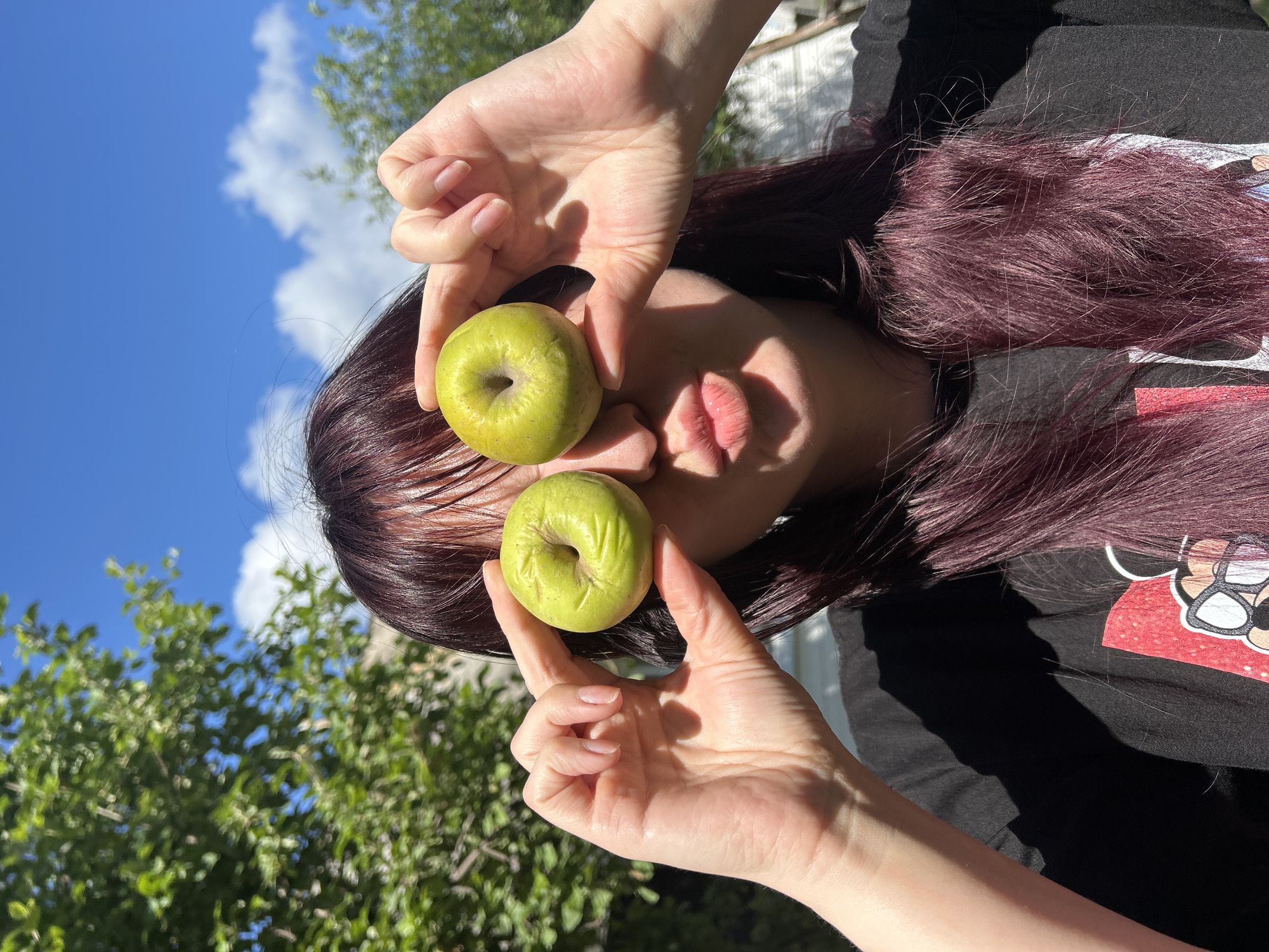 A young woman with dark, plum-colored hair is outdoors, holding two small green fruits in front of her eyes. She is puckering her lips as if for a kiss, and the sunlight is casting shadows across her face and black t-shirt. The background features a bright blue sky with scattered white clouds and lush green trees, suggesting a sunny day. The location is identified as the city of Foky, Russia.