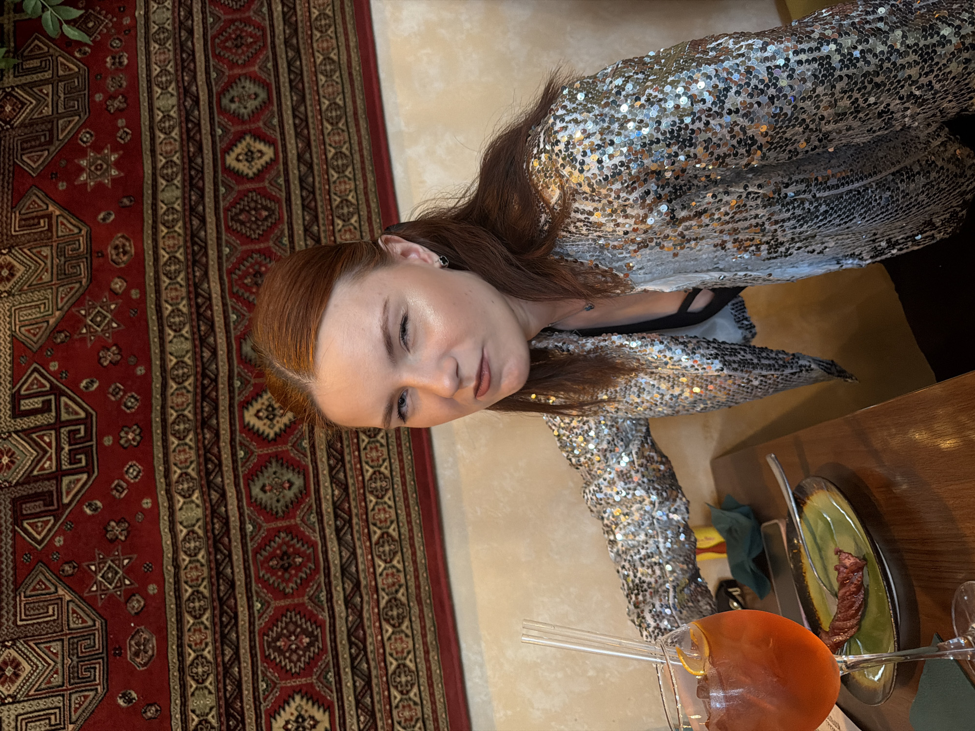 This image depicts a young woman in an indoor dining establishment, likely a restaurant or cafe, in Saint Petersburg, Russia. The woman, positioned in the mid-ground, has reddish-brown hair, styled partially pulled back, and fair skin with light makeup. She wears small, dark circular earrings and a sparkling silver sequined jacket over a dark top. Her expression is a wink with her right eye, her left eye is open, and her lips are slightly pursed, as she looks directly towards the viewer.
On the dark wooden table in the lower left foreground, a large stemmed glass contains an orange-colored cocktail, garnished with an orange slice and two clear straws. Adjacent to the glass, a green and dark-rimmed plate holds a single piece of cooked meat, resembling a grilled sausage or kebab, with a silver fork resting on it. A dark green napkin and other partially visible objects are also on the table. The background features a prominent red, black, and beige patterned tapestry or rug hanging on a light beige wall. The tapestry displays complex geometric patterns, star motifs, and interlocking designs, indicative of Central Asian or Caucasian textile art. A small portion of a green plant is visible in the upper right corner of the background.