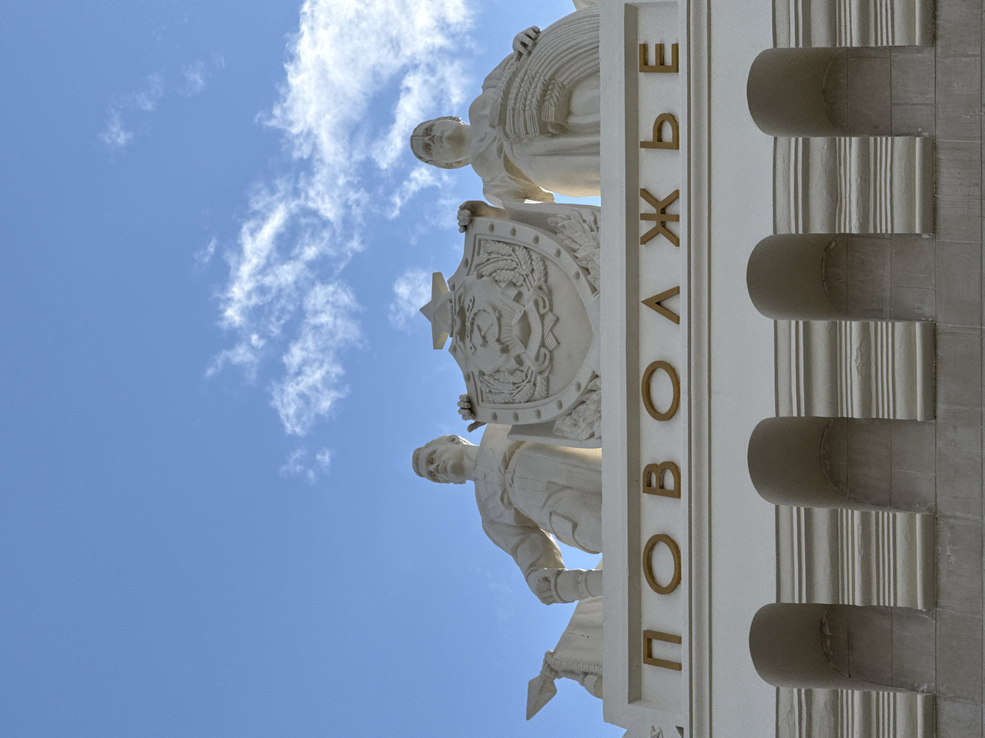This is a low-angle shot of a section of the facade of a building, likely an exhibition pavilion, in Moscow, Russia. The image is dominated by a bright blue sky with scattered white, wispy clouds, suggesting a clear day with moderate cloud cover. The lighting indicates it is daytime.
The main subjects are several ornate sculptural elements, rendered in a light-colored material, possibly stone or plaster. The sculptures depict figures and symbolic emblems. In the upper part of the image, a female figure holding a sheaf of wheat can be seen. Below her, a shield bearing a star and what appears to be a hammer and sickle is supported by another sculpted figure, a man in historical attire. The style of the sculptures is characteristic of Soviet-era public art, often found on buildings in Moscow, particularly at sites like VDNKh (Exhibition of Achievements of National Economy).
To the right of the sculptures, the building's facade features large, vertically fluted pilasters with rounded capitals, adding to the monumental and classical aesthetic. Along the edge of the facade, prominent gold-colored letters form Cyrillic text. The visible letters spell out "НОВ • О • ВОЛЖЬЕ" which likely signifies "New Volga" or a similar geographical or thematic reference. The overall impression is one of grandeur and civic pride, typical of the architecture from the Soviet period. The rotation of the image suggests it was taken with the camera tilted upwards, emphasizing the scale of the architecture against the sky.