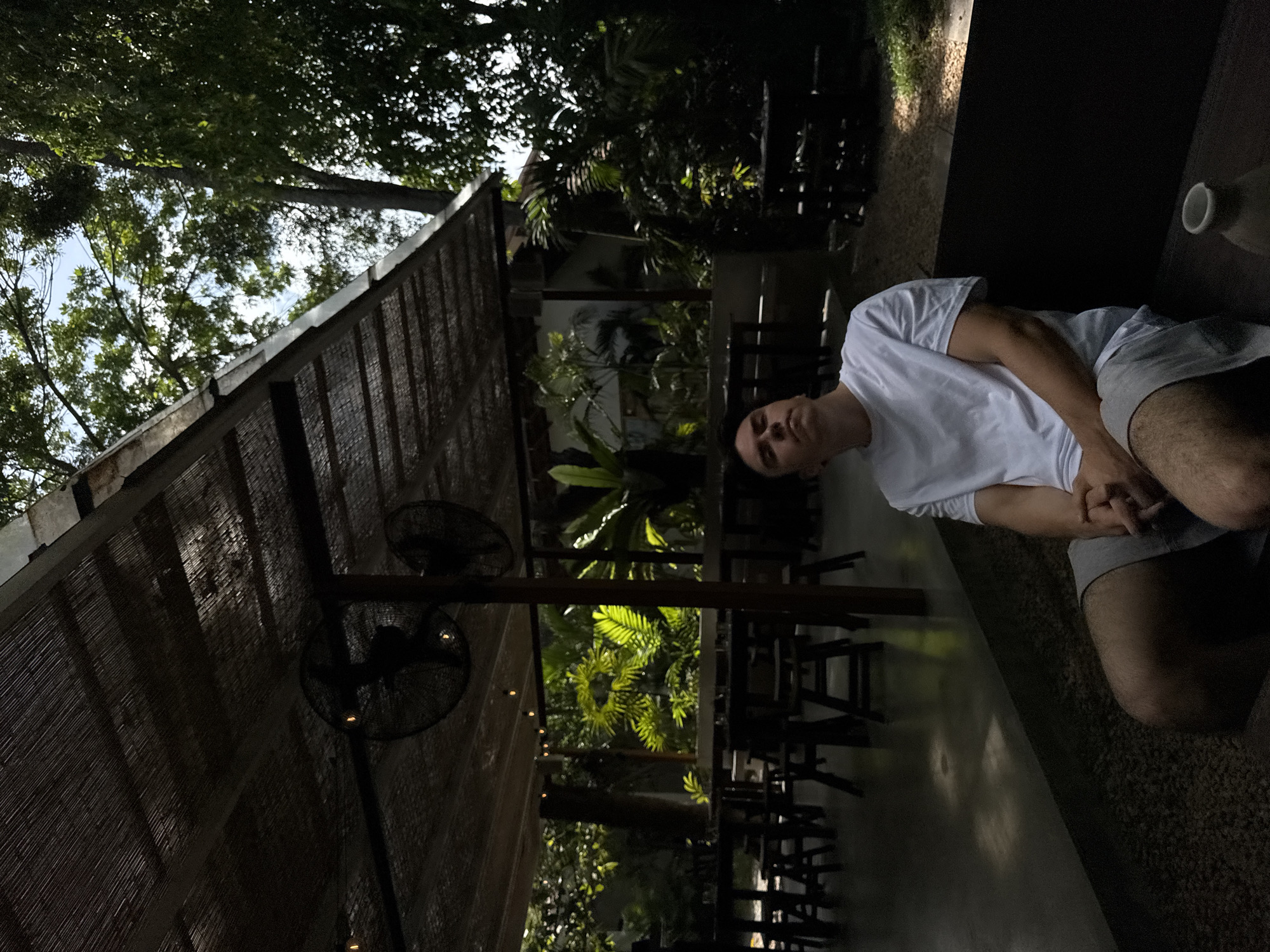 In Ahangama, Sri Lanka, a young man in a white t-shirt and grey shorts sits on a dark-colored bench, looking upwards and to his left. He is positioned in the foreground, with his hands clasped in his lap. Behind him, an outdoor seating area features multiple wooden tables and stools, partially obscured by the shadows cast by a woven roof overhead. Two large, black fans are suspended from the ceiling, and several small lights are visible. Lush green foliage and trees form the backdrop, with sunlight filtering through the leaves, creating dappled light on the ground. The overall setting appears to be a relaxed, open-air cafe or restaurant.