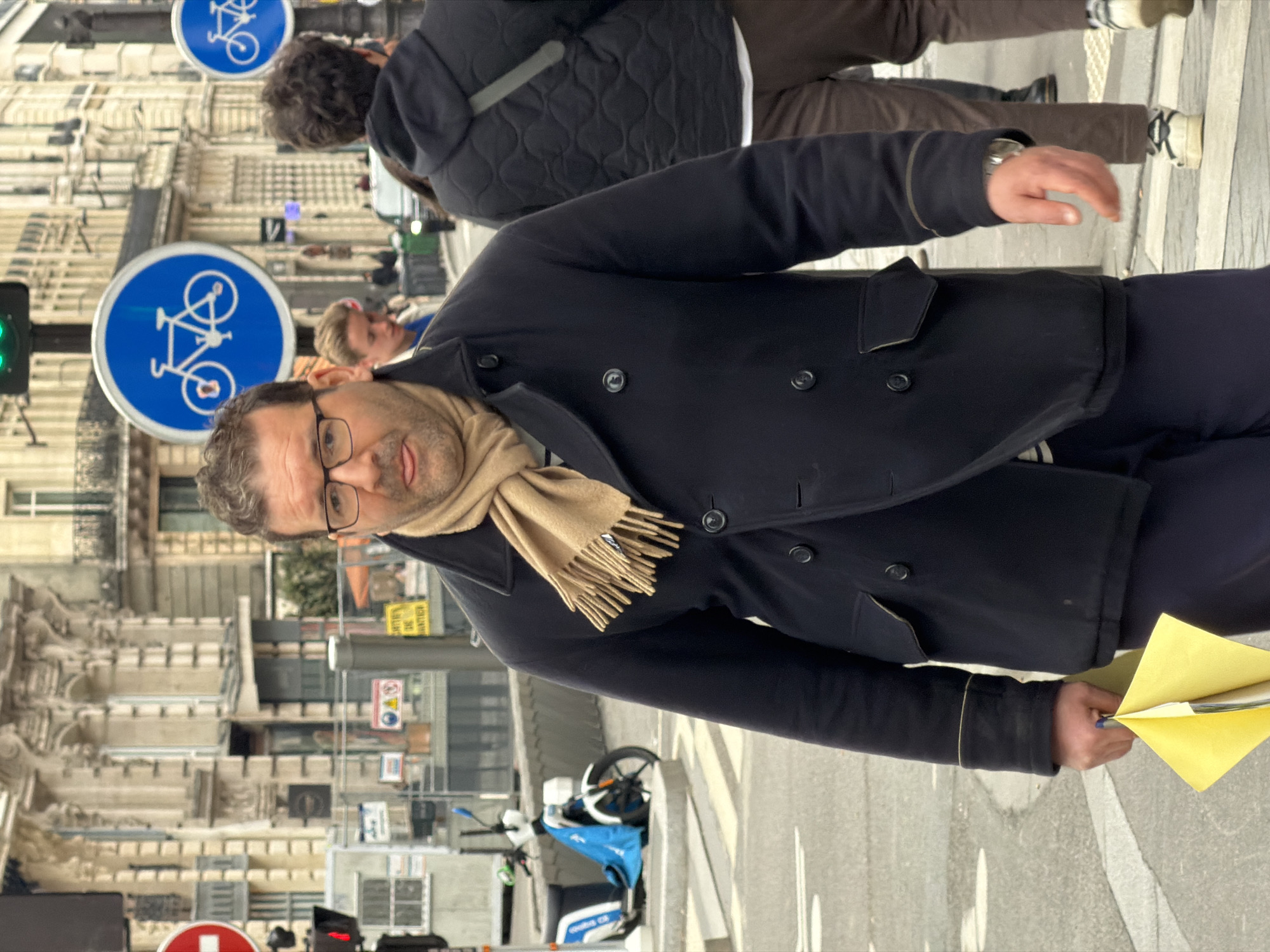 The scene depicts an urban streetscape in Paris, France, characterized by multi-story Haussmannian buildings with ornate limestone facades lining the background.
In the foreground, a middle-aged male, with short, dark, curly hair, a short beard, and black-framed glasses, walks towards the viewer, slightly right of center. He wears a dark navy double-breasted peacoat over a light tan fringed scarf and dark trousers. His right hand holds a folded piece of yellow paper, while a silver-toned watch is visible on his left wrist. He looks directly forward with a slightly pursed expression.
To the immediate right of the main subject, a second male figure is seen from behind, walking away. He has dark, curly hair and wears a black quilted jacket with a diagonal grey stripe on the back, brown trousers, and white sneakers. Further in the background, to the left of the main subject, a young individual with light hair is partially visible.
The environment includes a paved street with white pedestrian crossing stripes. Traffic infrastructure consists of a green traffic light and a red circular "no entry" sign on poles, alongside at least two prominent blue circular signs with white bicycle symbols, indicating a bicycle route or lane. Construction fencing, adorned with yellow warning signs reading "CHANTIER INTERDIT DE MONTER" and other cautionary symbols, demarcates a construction zone in the mid-ground. A white and blue shared scooter is parked on the sidewalk to the left. The overall lighting suggests an overcast or muted daytime.