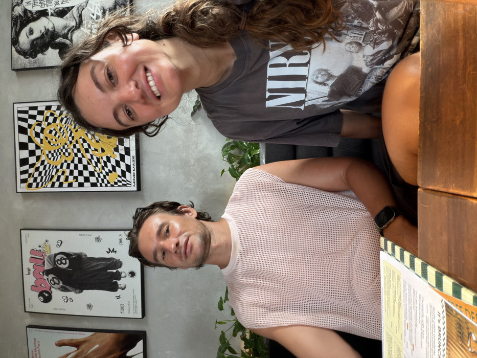 A smiling woman in a Nirvana shirt and a man in a pink mesh tank top pose for a photo. The woman is leaning in from the top left of the frame. Behind them are various framed artworks, including a checkerboard design and one resembling a vintage movie poster. A small potted plant is visible between the people. The man is wearing a smartwatch. A menu rests on a wooden table to his right.