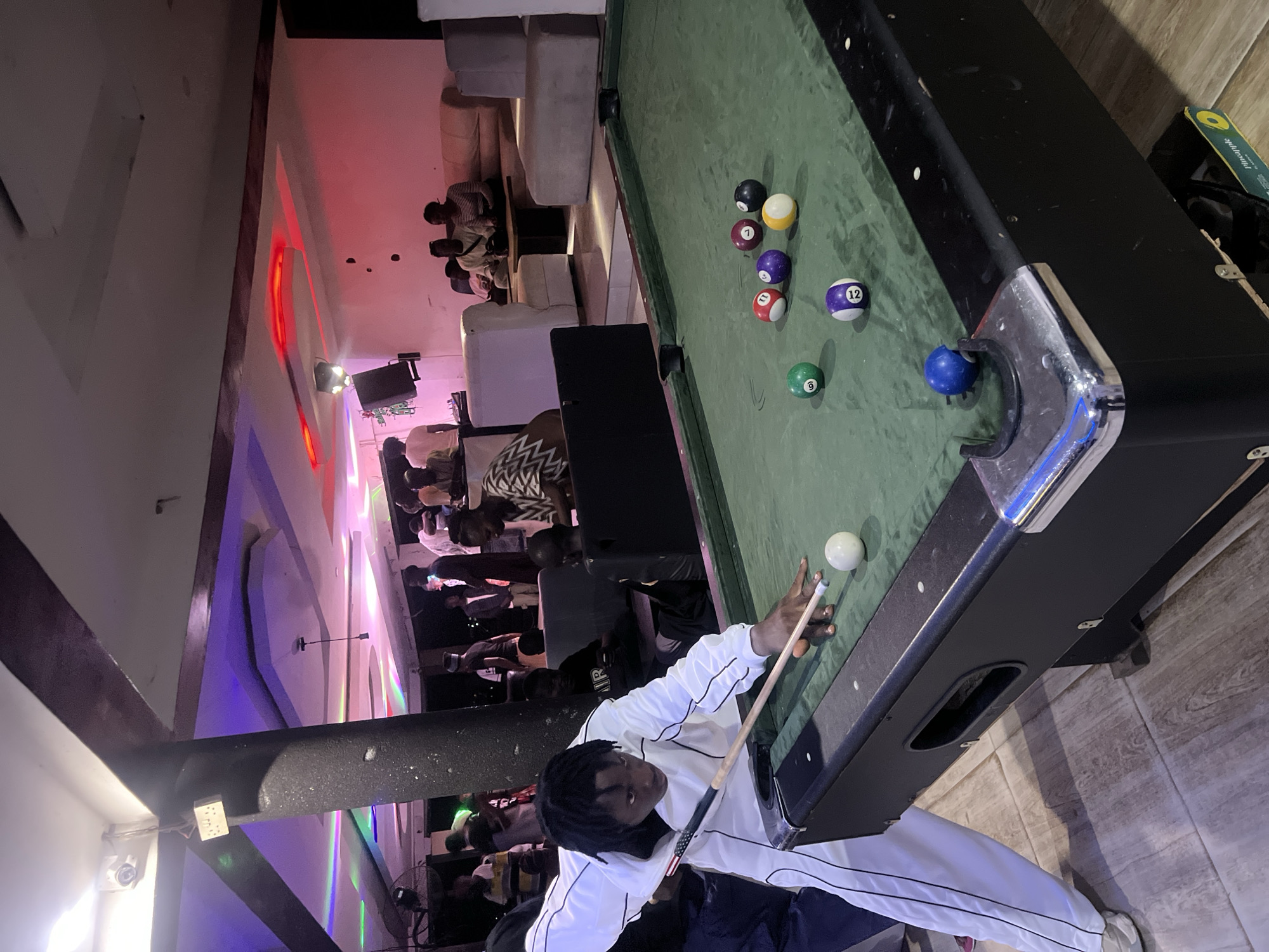 The scene is a bar in Howayi, Nigeria, illuminated with colorful neon lights and filled with patrons. A man with dark hair styled in dreadlocks, wearing a white tracksuit, is playing pool. He is holding a pool cue and aiming at a white ball on a green pool table. Several other pool balls are scattered on the table, including black, yellow, purple, red, and green. Behind him, other people are seated at tables or standing, and some are wearing patterned clothing. Couches are pushed against the wall in the distance.