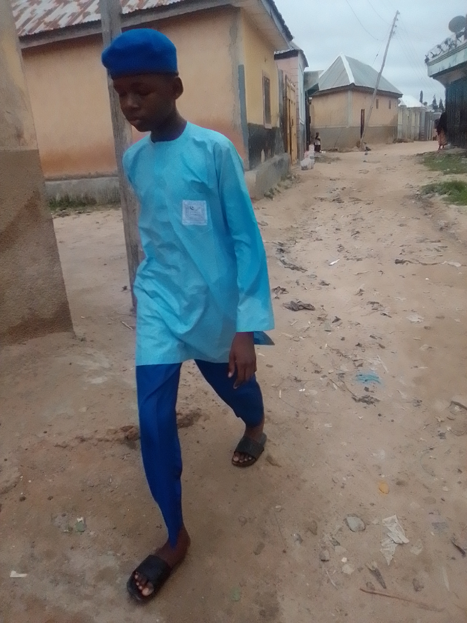 A young boy, appearing to be of African descent, stands in a dirt alleyway in Gombe, Nigeria. He is wearing a vibrant blue cap and a matching light blue, long-sleeved tunic with a small white emblem on the left chest. His trousers are a darker shade of blue, and he wears black sandals. The boy is looking down and to his left, with his body angled towards the viewer.
The environment is an unpaved alley lined with buildings. To the left, a building with a textured, light brown wall is partially visible. To the right and further down the alley, other structures with corrugated metal roofs and light-colored walls are present, some with simple doorways. The ground is covered in dry, sandy dirt, with some scattered debris, including small pieces of plastic and dried vegetation. Overhead, the sky is overcast, suggesting a cloudy or hazy day. The overall impression is that of a typical residential street in a developing urban area.