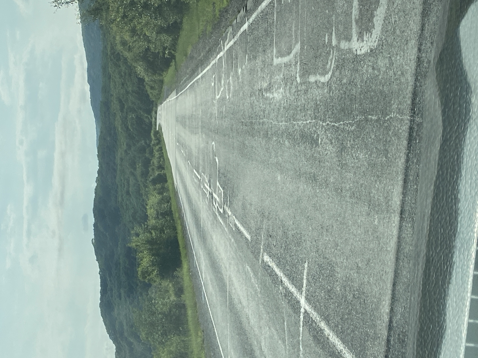An eye-level, medium shot captures a highway in Kravtsovka, Russia, on a cloudy day. The asphalt road, marked with white lines, stretches into the distance, curving slightly to the left. The road surface appears worn, with some patches of repair or damage. On the right side of the road, a white line delineates the shoulder, beyond which is a grassy verge and dense green trees. The left side of the road also has a grassy verge and trees. In the background, rolling hills covered in lush green forest stretch towards a hazy horizon under a pale blue sky with scattered clouds. The foreground shows the edge of a vehicle's dashboard or interior, rendered in a metallic, textured gray.