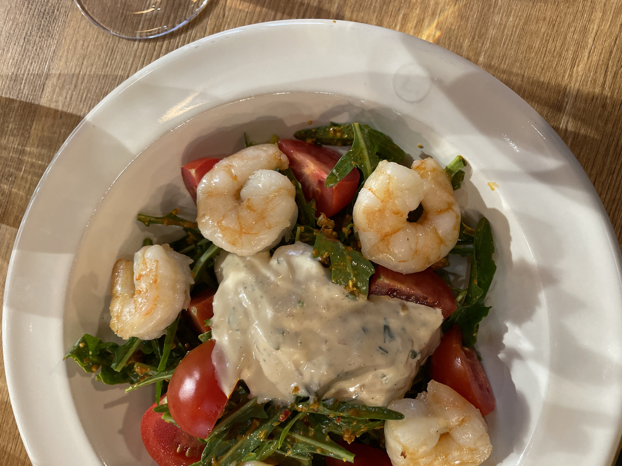 The image shows a white, shallow bowl filled with a salad. The salad consists of cooked shrimp, halved red cherry tomatoes, and green leafy vegetables, possibly arugula. A creamy, light-colored sauce is generously dolloped on top of the salad. The bowl sits on a wooden surface, possibly a table, with a small portion of a glass visible in the upper left corner. There is no clear indication of the location being Tbilisi, Georgia, nor are there any people visible in the image. The focus is solely on the presentation of the salad within the bowl.