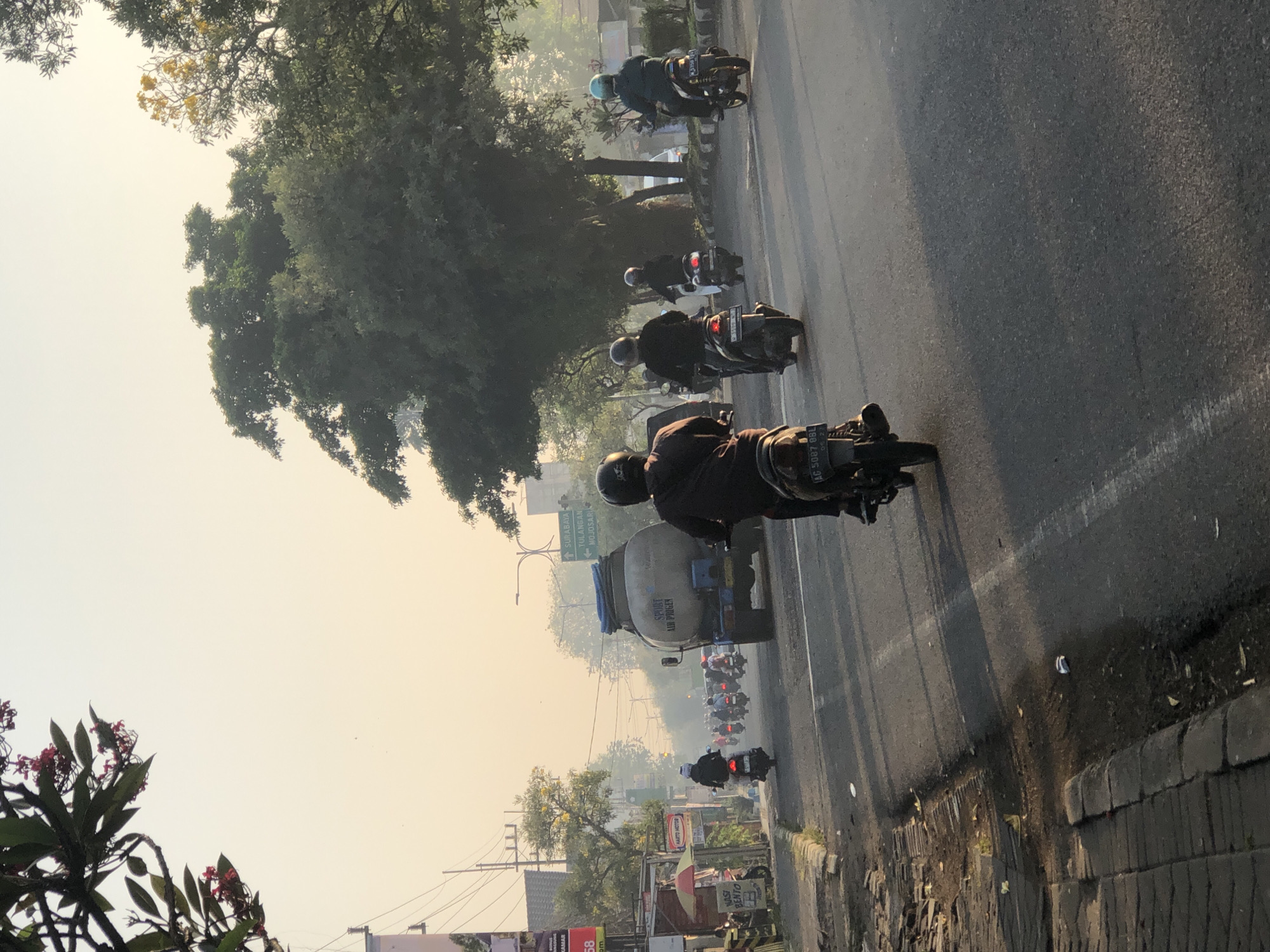 This is a photograph taken on a street in Kludan, Indonesia, likely during the morning given the soft, hazy sunlight and the presence of mist or fog in the distance. The scene depicts a busy road with numerous motorbikes navigating the traffic.
In the foreground, a person on a motorbike is captured from behind, wearing a dark jacket and helmet. To their left, another person on a motorbike also faces away from the camera. Further ahead, a line of motorbikes and riders are moving along the road, some with helmets and others with headscarves. A large tanker truck, labeled "SPORT AIR PRIGER," is a prominent object in the middle of the road, also moving with the traffic. On the left side of the road, there are buildings and storefronts, including one with an umbrella and a sign that reads "NASI BENTO". Trees line the right side of the road, casting shadows on the asphalt. Overhead, telephone wires are visible. The overall impression is of a typical, bustling urban or suburban street scene in Indonesia during the early part of the day.