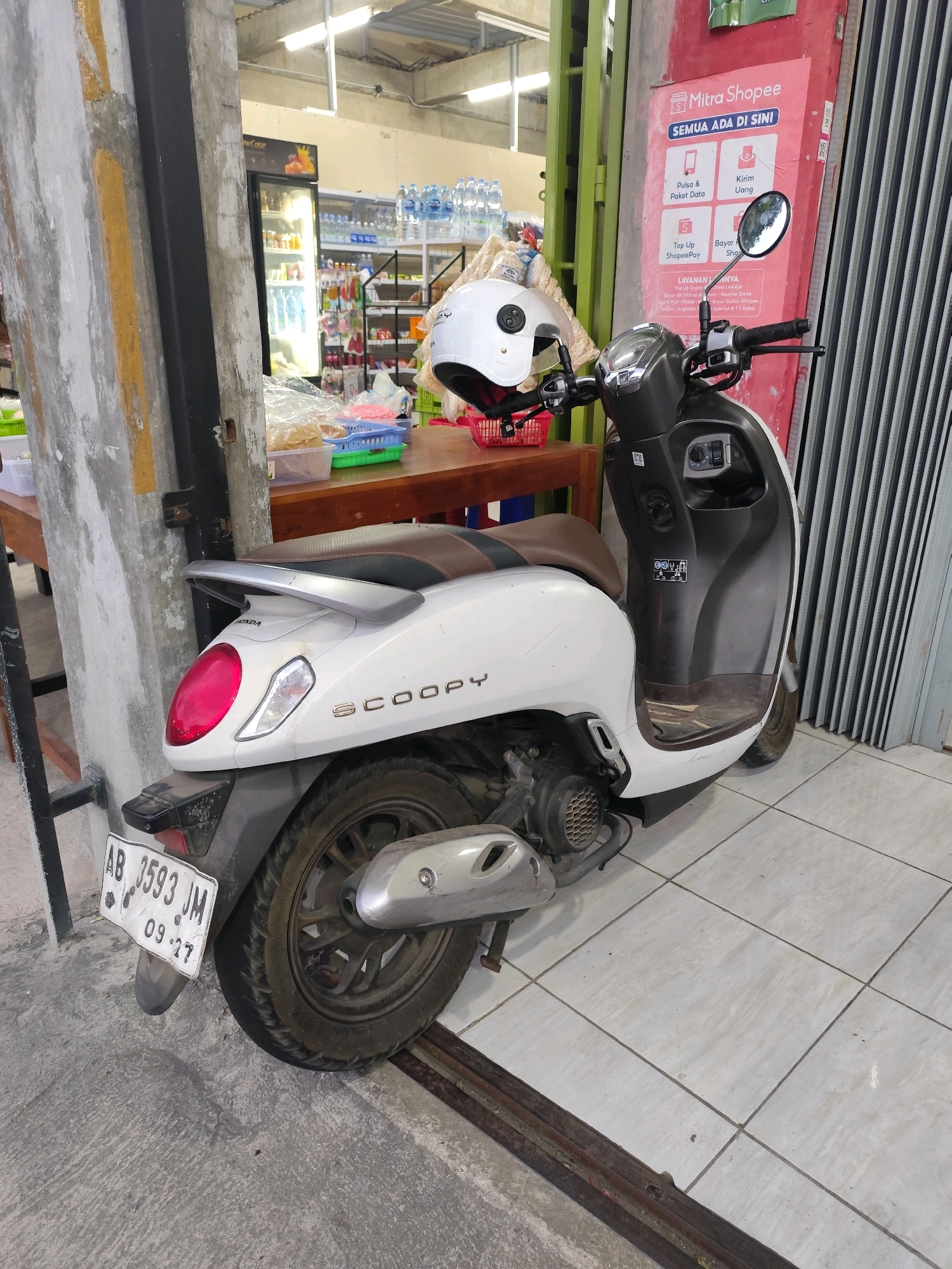 A white Honda Scoopy motorcycle is parked outside a convenience store in Bambang Lipuro, Indonesia. The motorcycle is facing right and is parked on a tiled floor. A white helmet is placed on the handlebars. The store has a red exterior with a Shopee Mitra advertisement displayed on the wall. Inside the store, shelves are stocked with bottled water and various products. The license plate of the motorcycle reads "AB 3593 JM 09-27". The scene appears to be in daylight, with artificial lighting visible inside the store.